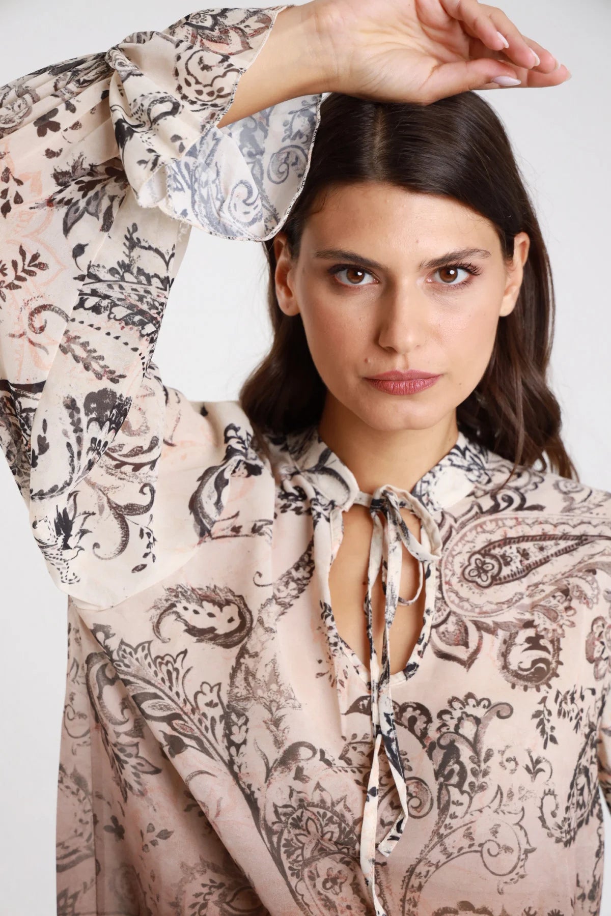 Blusa fantasia paisley baroque chiffon panna ecru cioccolato moro degrade dettaglio frontale busto mano testa scollo V laccetto fiocco colletto drappeggiante manica balloon bishop polsino balze ruches svasato laccetto fiocco dettaglio polsino - modella frontale busto mano testa sguardo frontale intenso capelli castani lisci sfondo bianco