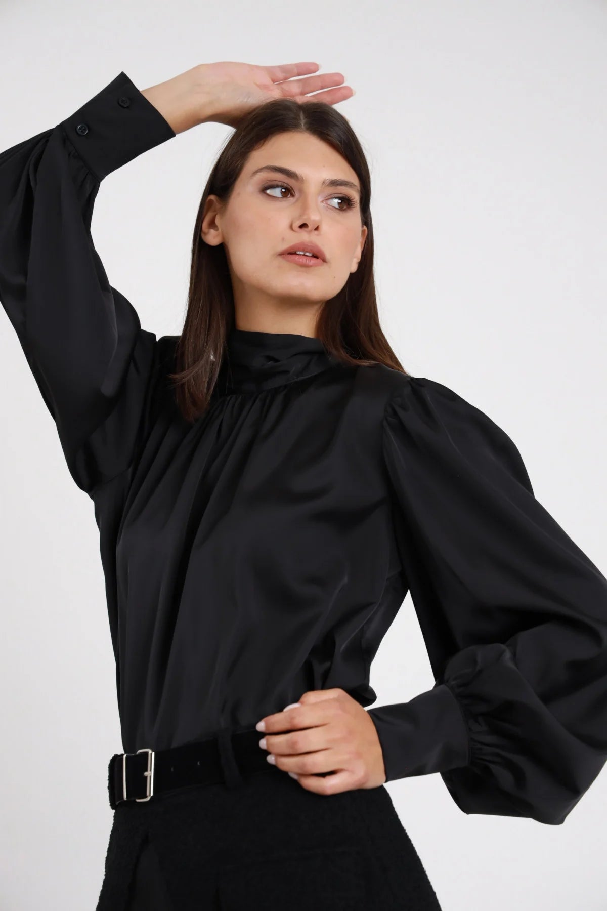 Camicia Fiocco Retro nera 100% poliestere satin lucido cowl neck drappeggiato polsino bottone nero spalle puff manica lunga balloon frontale busto 3/4 mano fronte sguardo laterale alto - modella busto 3/4 mano fronte sguardo laterale alto capelli castani scuri lisci sfondo bianco gonna costine nera cintura nera fibbia argento