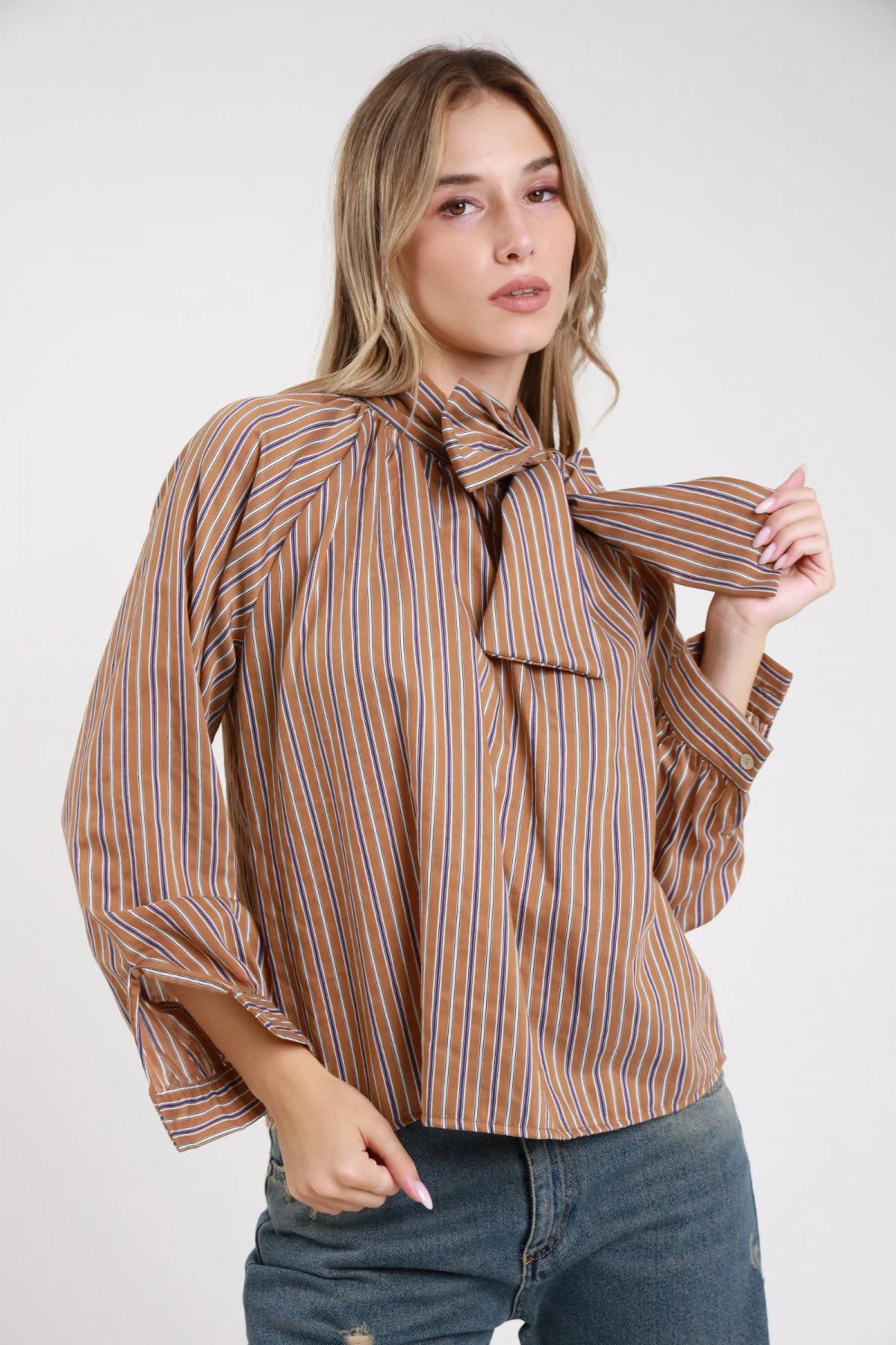 Camicia righe fiocco lavallière cammello blu arancio ampia - modella busto tre quarti frontale mano al fiocco jeans chiari capelli biondi mossi sguardo laterale