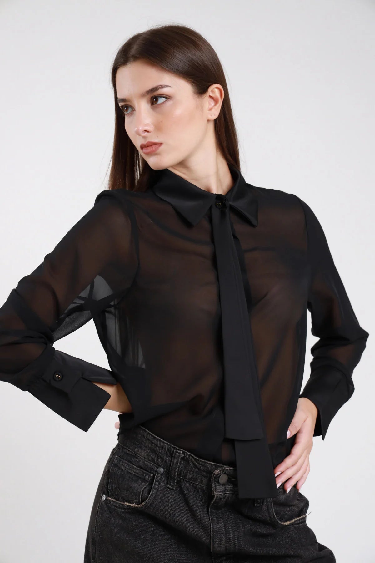 Camicia chiffon trasparente nera cravatta lavallière nera satin colletto classico bottone gioiello cristallo manica lunga polsino doppio oversize frontale busto mani fianchi - modella frontale mani fianchi sguardo laterale capelli castani lisci sfondo bianco jeans denim nero