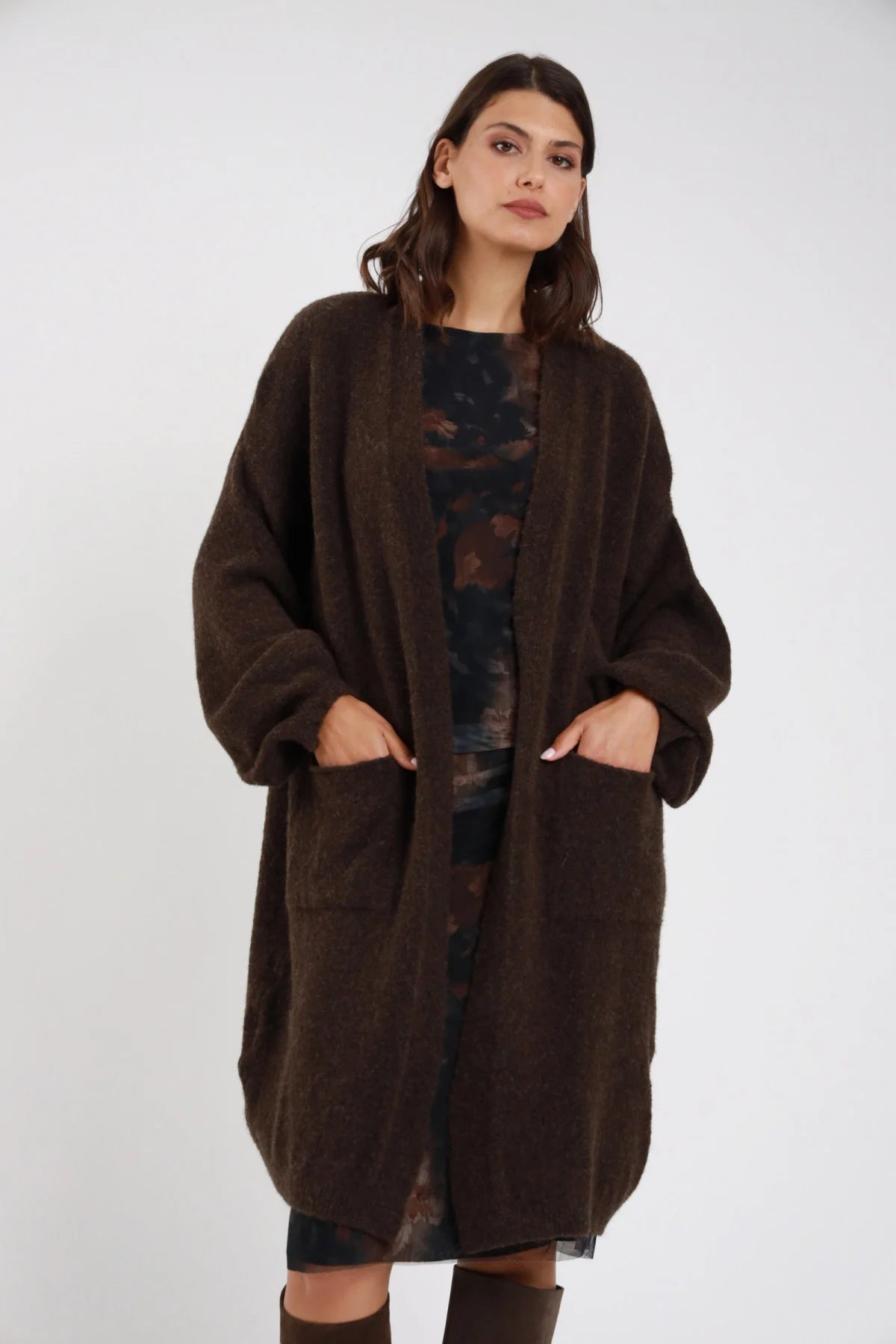 Cardigan lungo midi maglia bouclé eyelash marrone cioccolato fondente aperto scollo V revers batwing oversize cocoon tasche patch frontale intera mani tasche - modella frontale intera mani tasche sguardo frontale capelli castani ondulati sfondo bianco stivali tacco marroni abito fantasia floreale scuro