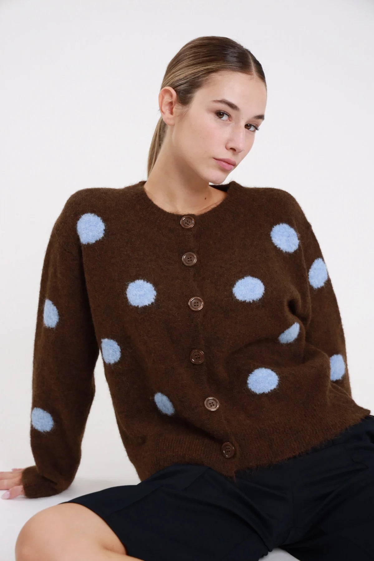 Cardigan pois cioccolato moro azzurro baby fluffy eyelash frontale seduta tre quarti maxi pois jacquard tridimensionali azzurro girocollo abbottonatura bottoni cioccolato corpo boxy - modella frontale seduta tre quarti sguardo laterale pantaloni neri capelli castani raccolti