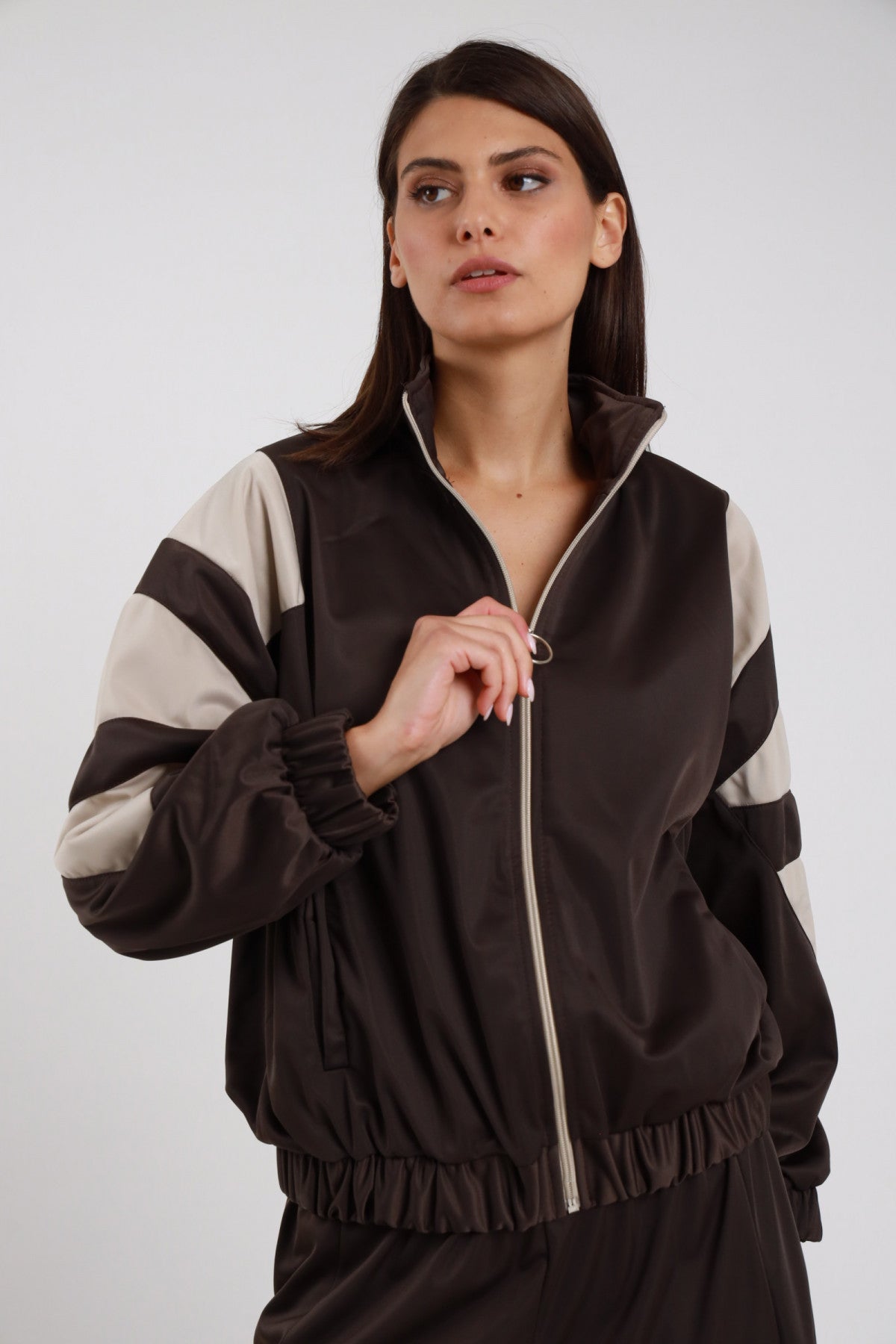 Giacca Zip Bande Diagonali marrone cioccolato scuro raso satin collo alto rialzato bande diagonali panna sabbia larghe obliquo maniche zip anello argento elastico fondo bomber crop - modella busto 3/4 frontale posa mano zip sguardo laterale alto capelli castani lisci sfondo bianco pantalone palazzo marrone coordinato