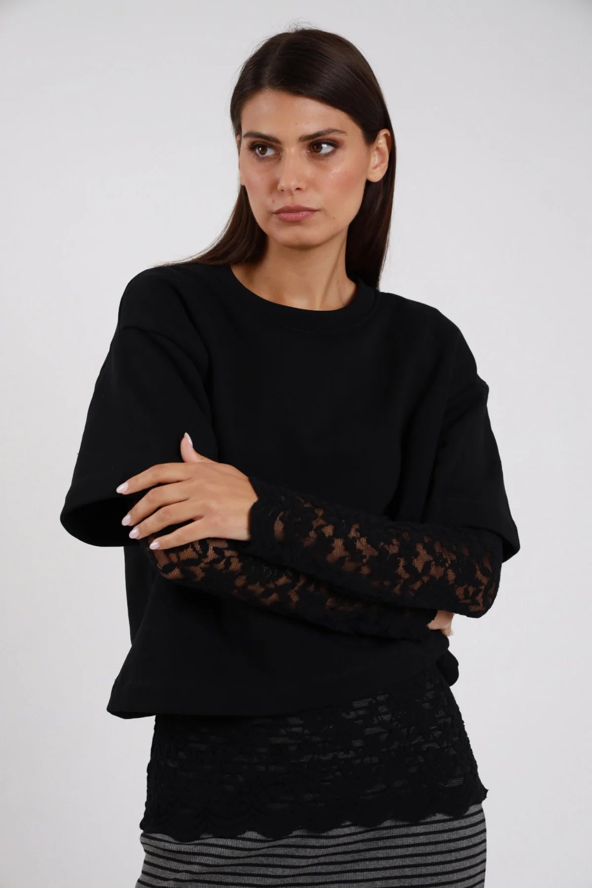 Felpa Girocollo Oversize Inserto Pizzo Floreale nera 100% cotone girocollo costina manica 3/4 inserto pizzo floreale nero polso avambraccio orlo pizzo smerlato silhouette boxy - modella busto 3/4 frontale braccia incrociate sguardo laterale capelli castani lisci sfondo bianco gonna a righe grigio bianco