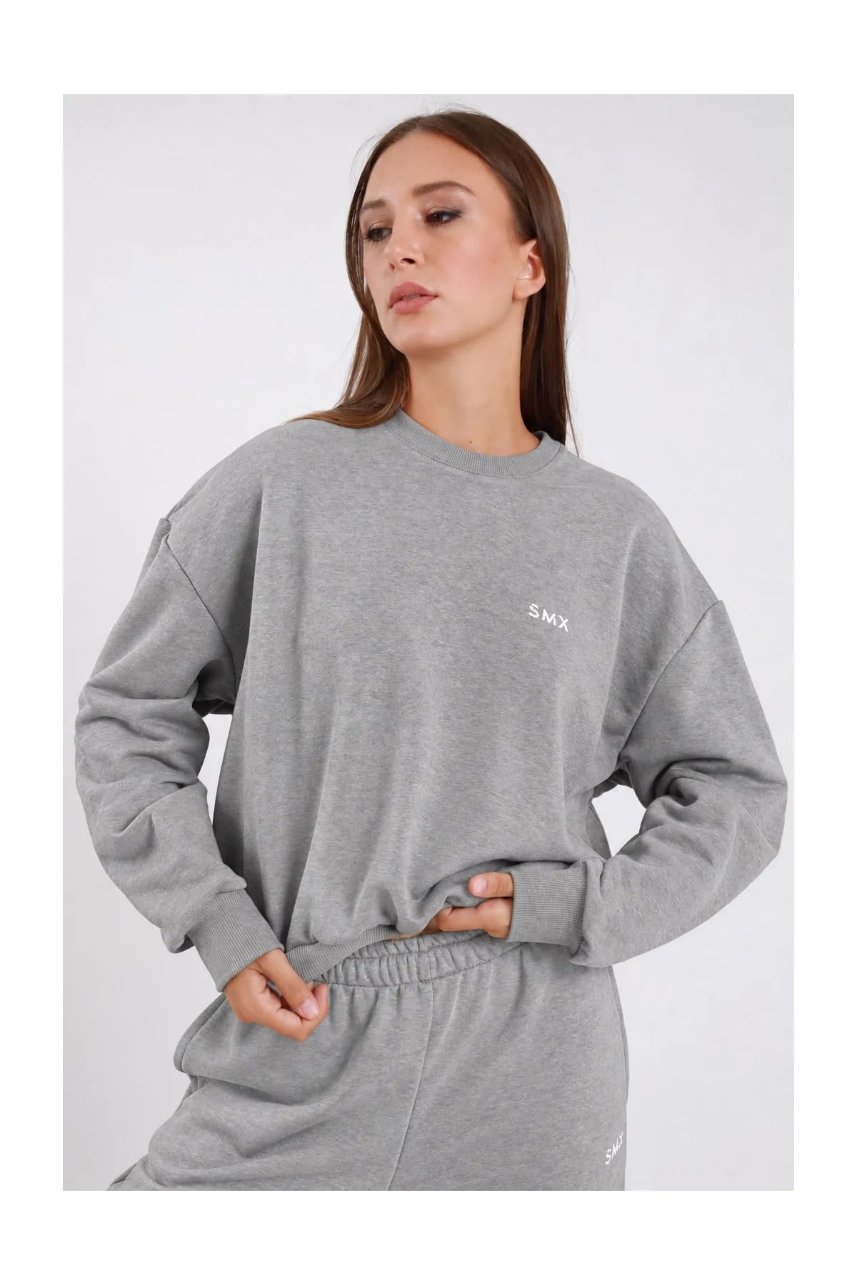 Felpa Girocollo DAYTHRU grigio melange chiaro logo SMX ricamato bianco petto cuciture esterne decorative a vista manica oversize polsino costine boxy busto frontale 3/4 mani vita sguardo basso - modella busto frontale 3/4 mani vita sguardo basso capelli castani lisci sfondo bianco pantalone tuta jogger grigio melange coordinato