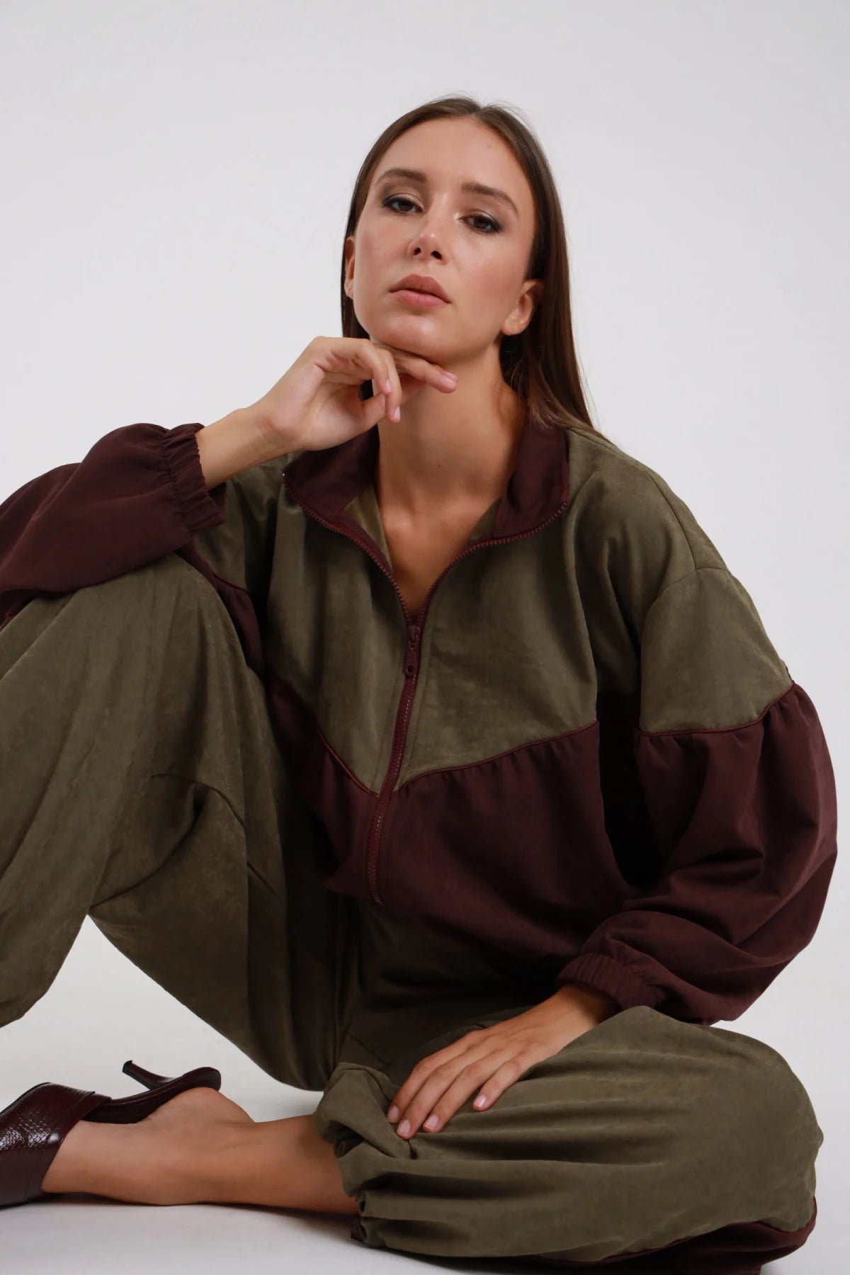 Bomber Bicolor verde oliva bordeaux colletto alto zip aperta maniche balloon bordeaux silhouette completa seduta posa editoriale mano mento - modella seduta posa editoriale mano mento sguardo laterale capelli castani lisci sfondo bianco pantaloni verde oliva décolleté marroni tacco kitten
