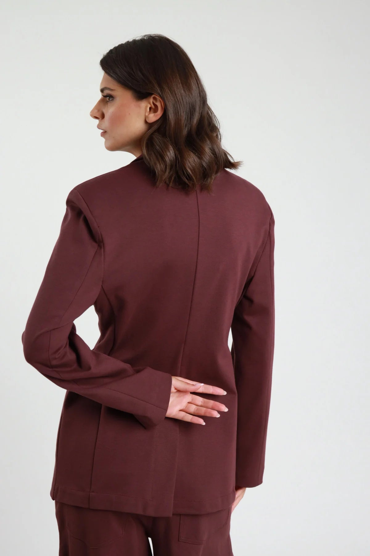 Blazer Punto Milano bordeaux mogano revers V profondo bottone dorato decorativo tasche filetto patta spalle strutturate retro intera mano schiena - modella retro intera mano schiena sguardo laterale capelli castani scuri sfondo bianco pantalone uovo bordeaux