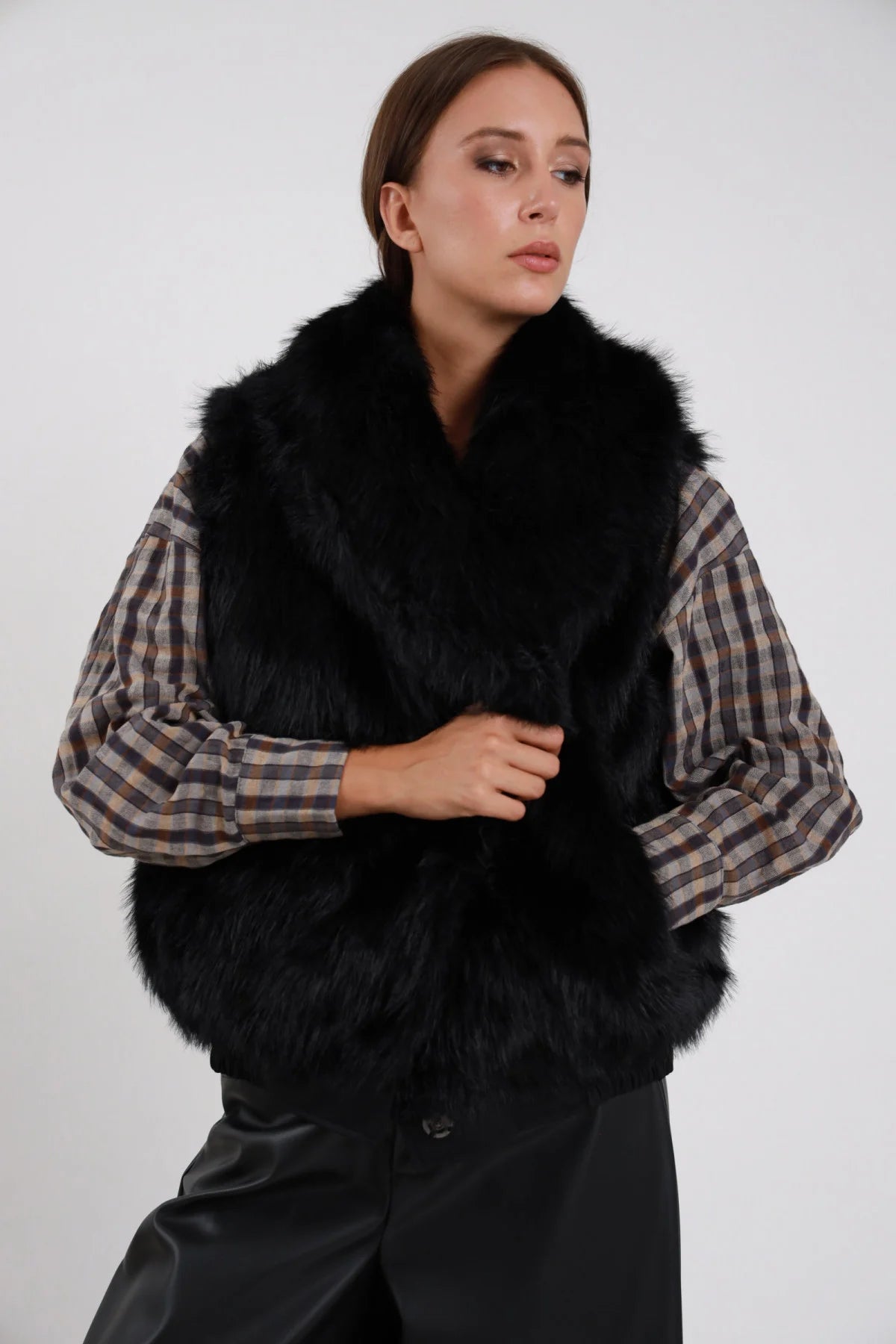 Gilet Ecopelliccia nero full ecopelliccia nera integrale colletto V senza maniche busto frontale 3/4 mano chiusura sguardo laterale - modella busto frontale 3/4 mano chiusura sguardo laterale capelli castani lisci sfondo bianco camicia scozzese beige grigia pantaloni neri ecopelle