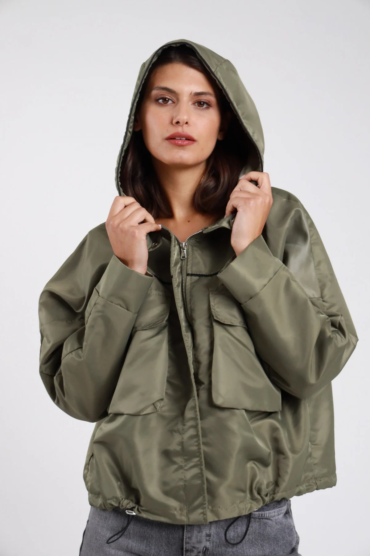Giacca giubbino nylon tecnico verde militare kaki olive impermeabile cappuccio ampio alzato regolabile bottoni pressione snap argentati tasche cargo maxi frontali pattina snap zip frontale aperta manica balloon raglan polsino elastico coulisse corpo oversize boxy vita hip fondo coulisse - modella frontale intera cappuccio alzato mani che regolano sguardo frontale intenso capelli castani corti mossi sfondo bianco jeans denim grigio