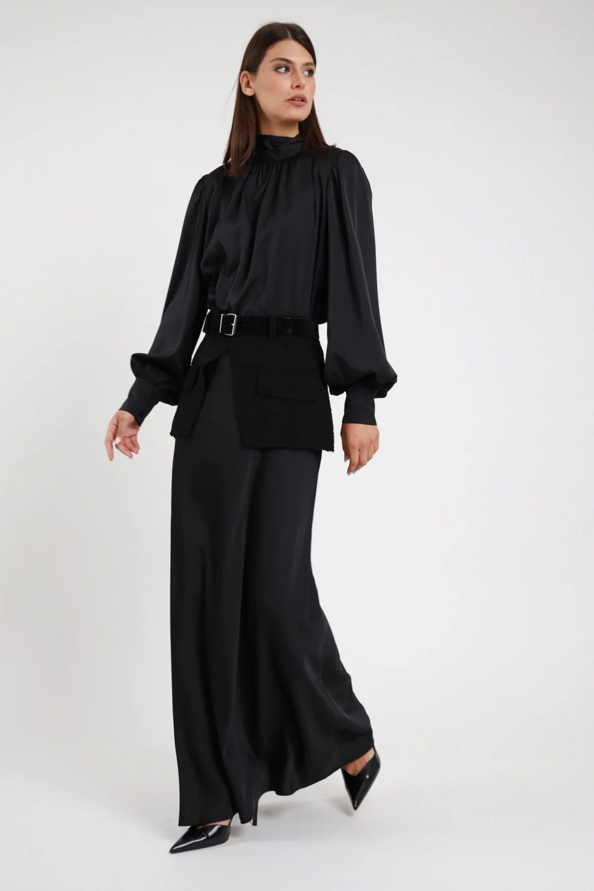 Gonna Lunga Cintura nera 100% poliestere satin svasata flared floor length fascia maglia cintura fibbia argento total look satin nero frontale intera 3/4 laterale posa in cammino - modella intera 3/4 laterale posa in cammino sguardo laterale capelli castani scuri lisci sfondo bianco total look camicia fiocco retro gonna satin nera décolleté neri punta
