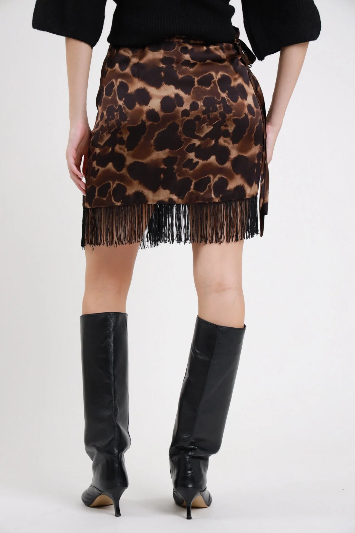 Minigonna Animalier Frange leopardata marrone beige nero 100% viscosa vita laccio cintura laterale annodato fiocco frange nere sull orlo posteriore vita gamba - modella vita gamba posteriore sfondo bianco maglia nera stivali ginocchio neri pelle