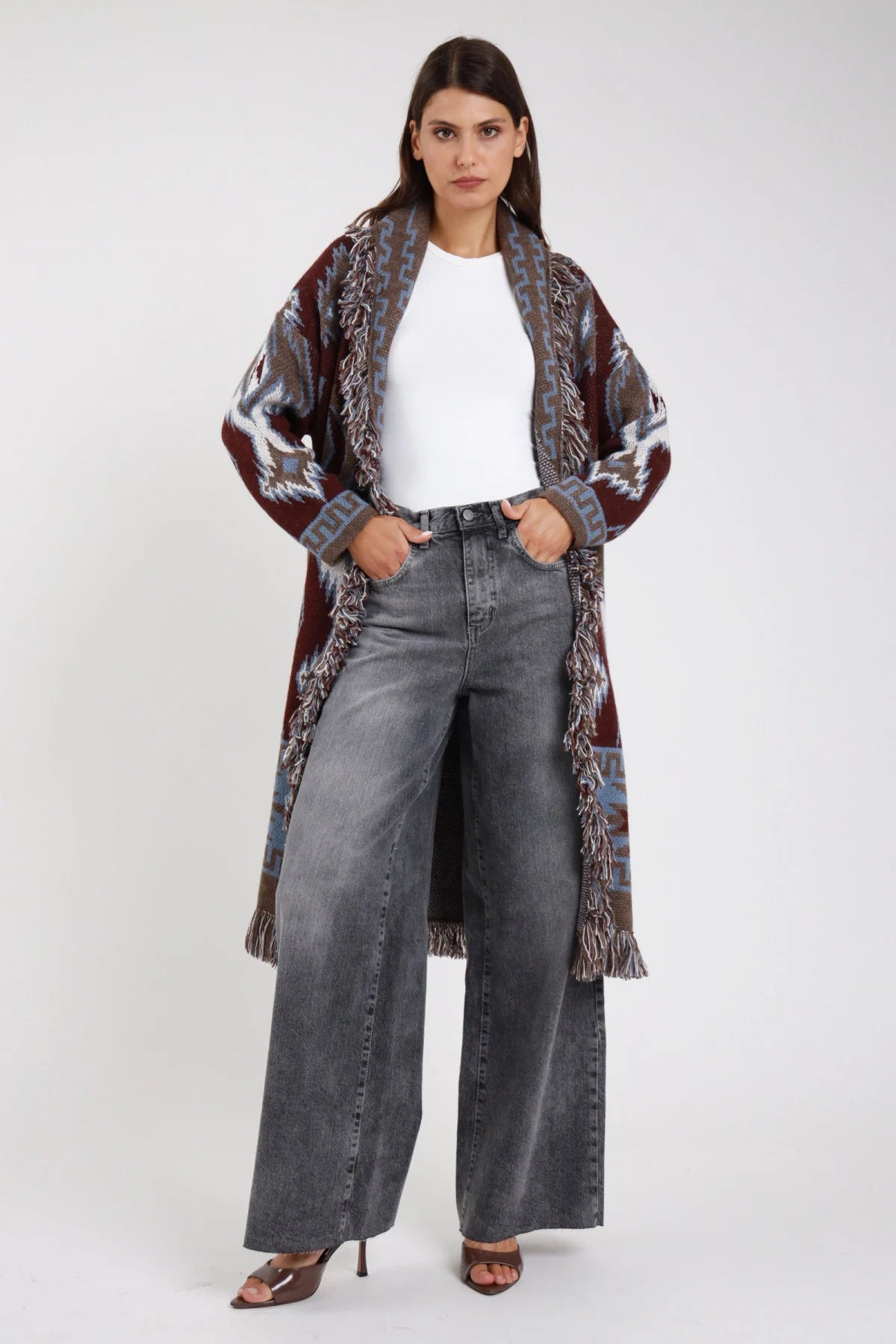 Jeans wide leg palazzo grigio stonewashed vita alta 5 tasche gamba larga maxi orlo grezzo frontale intera mani tasche con cardigan azteco bordeaux frange - modella frontale intera mani tasche sguardo frontale capelli castani scuri ondulati sfondo bianco cardigan azteco bordeaux frange t-shirt bianca sandali tacco