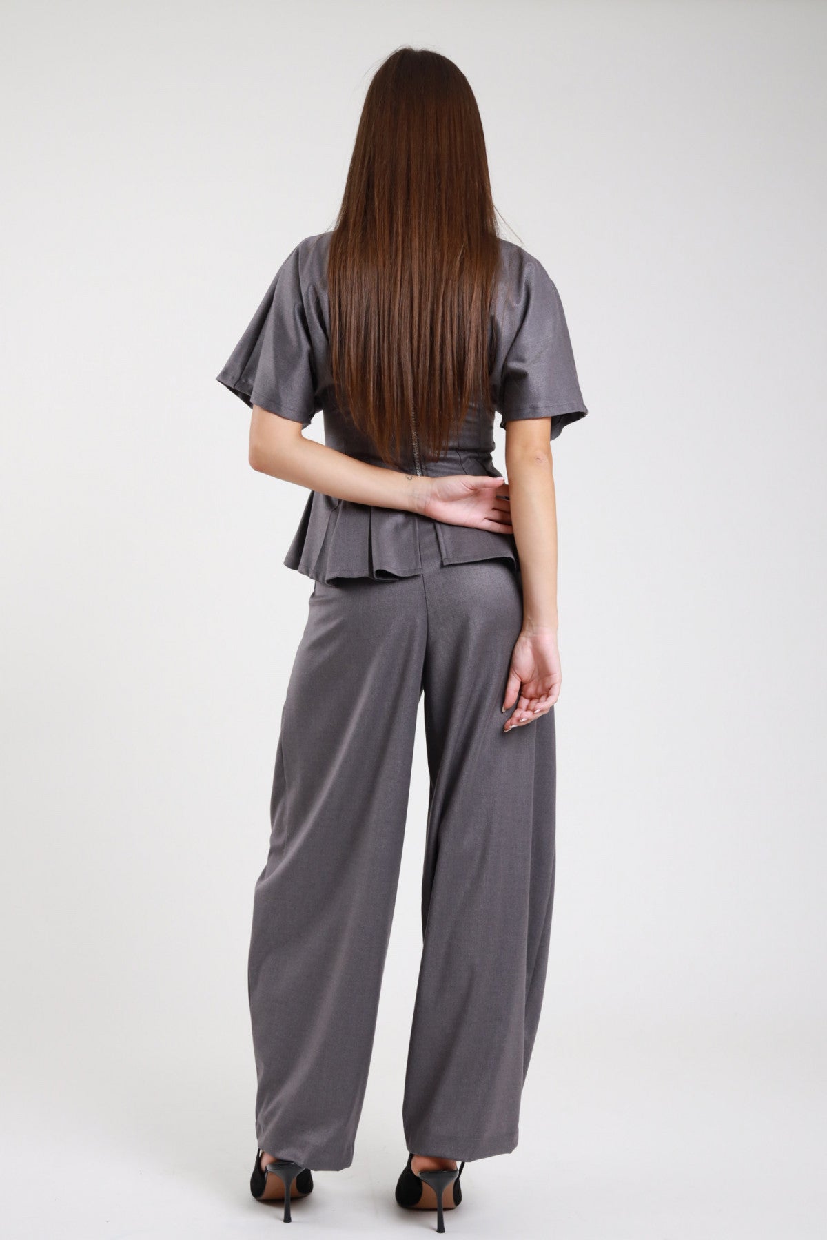 Pantaloni wide leg balloon grigio ardesia antracite crêpe stretch fascia elastica vita alta pince drappeggio gamba ampia pouching retro intera - modella retro intera mani intrecciate dietro schiena sguardo basso capelli castani scuri lisci lunghi sfondo bianco top maglia zip zip posteriore argento peplum grigio coordinato décolleté nero tacco