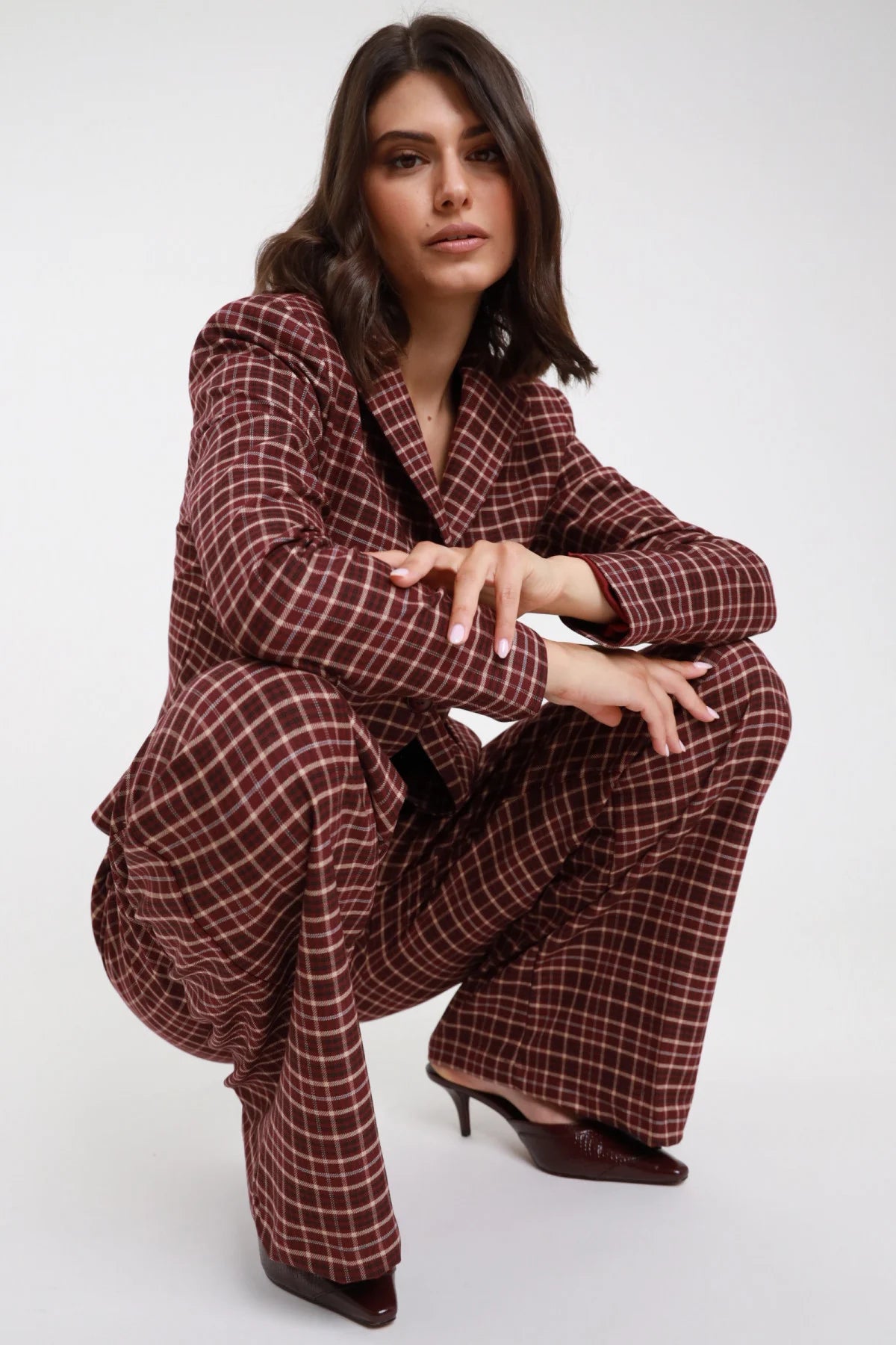 Pantalone Check tartan quadri bordeaux ruggine wide leg palazzo maxi vita elastico posa seduta accovacciata con Blazer Check bordeaux - modella seduta accovacciata frontale sguardo frontale capelli castani scuri sfondo bianco blazer check bordeaux kitten heel bordeaux vernice