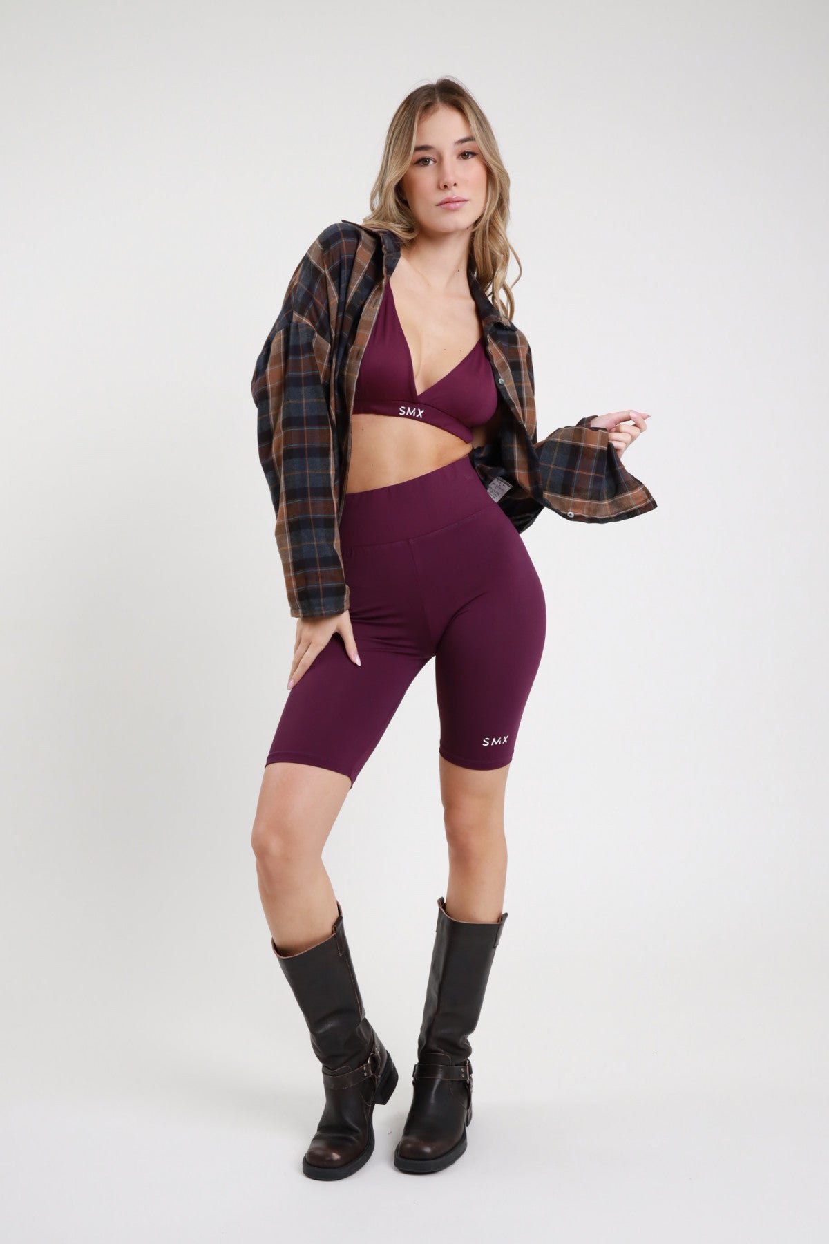 Short Ciclista SMX Limited Edition viola melanzana banda logo SMX vita alta logo SMX coscia laterale ben visibile lunghezza ginocchio body-fit frontale intera 3/4 - modella frontale 3/4 capelli biondi mossi sfondo bianco bralette triangolo viola SMX coordinata giacca tartan scozzese oversize aperta stivali neri alti abbinati