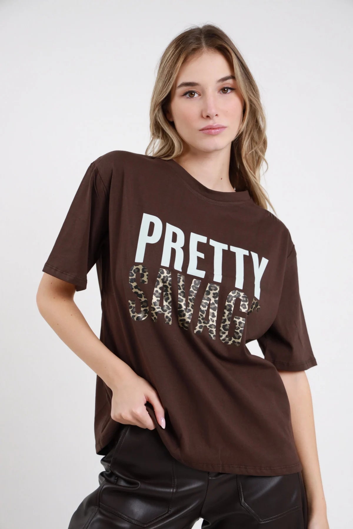 T-shirt Pretty Savage marrone cioccolato 100% cotone stampa PRETTY bianco acquamarina SAVAGE leopardato beige marrone girocollo manica corta silhouette oversize drop lunga - modella busto 3/4 frontale posa mano tasca sguardo frontale soft capelli biondi mossi sfondo bianco pantaloni ecopelle nera