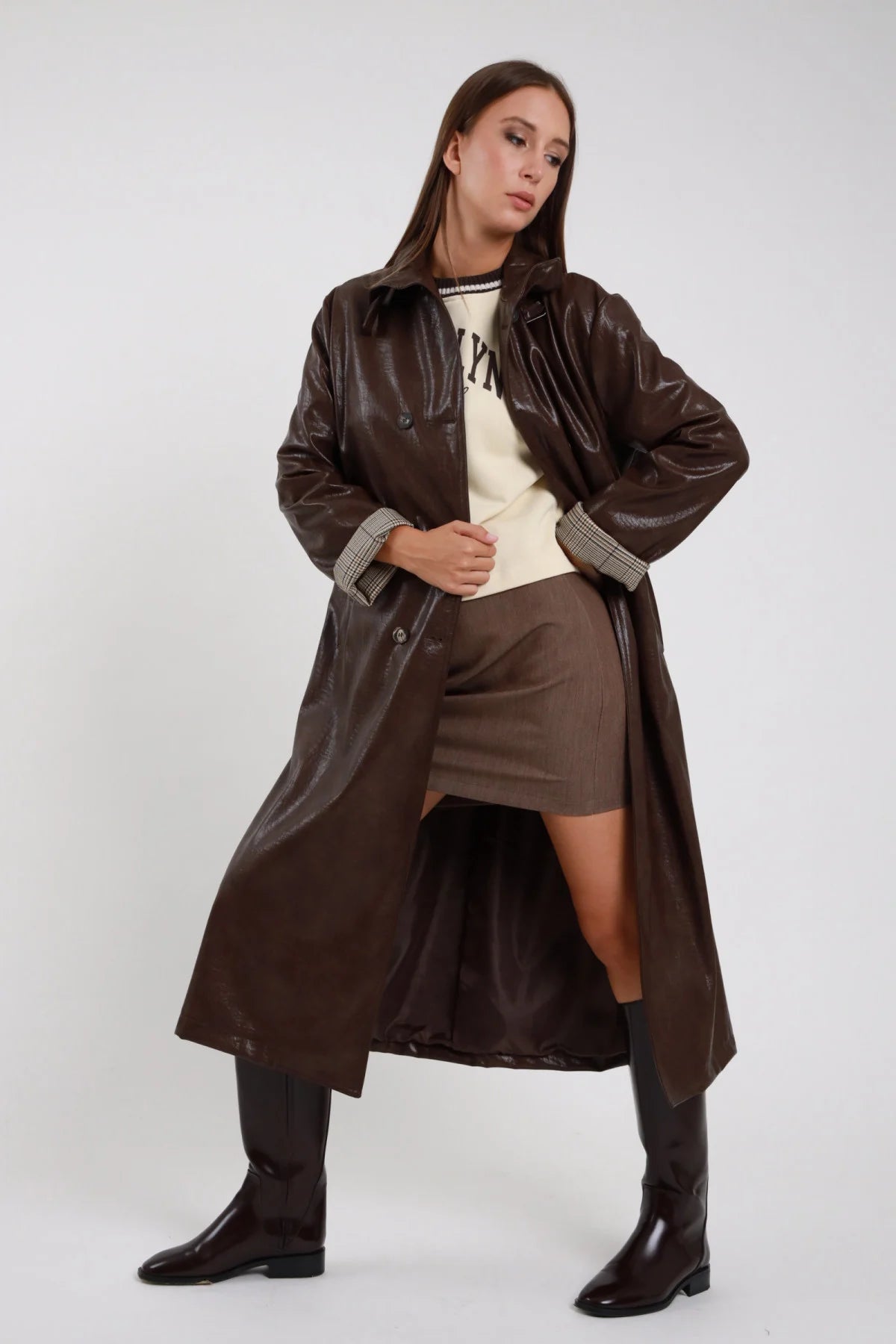 Trench Ecopelle marrone cioccolato scuro verniciata laccata crinkle doppiopetto tasche frontali polsino check silhouette maxi midi frontale intera posa dinamica gamba alzata aperto - modella frontale intera posa dinamica gamba alzata sguardo laterale capelli castani lisci sfondo bianco felpa panna brooklyn minigonna marrone stivali neri alti ginocchio piatto