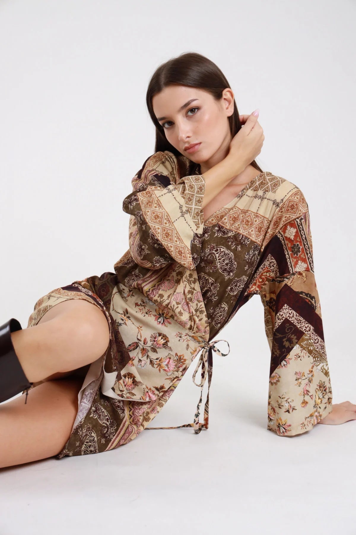 Abito kimono fantasia patchwork paisley baroque floral beige panna cioccolato moro frontale seduta terra sguardo laterale scollo V fiocco drappeggiante manica kimono batwing ultralarga polsino campana flare cintura laccetto nodo frontale orlo asimmetrico mini stivali neri - modella frontale seduta terra sguardo laterale intenso stivali neri al ginocchio capelli castani lisci sfondo bianco