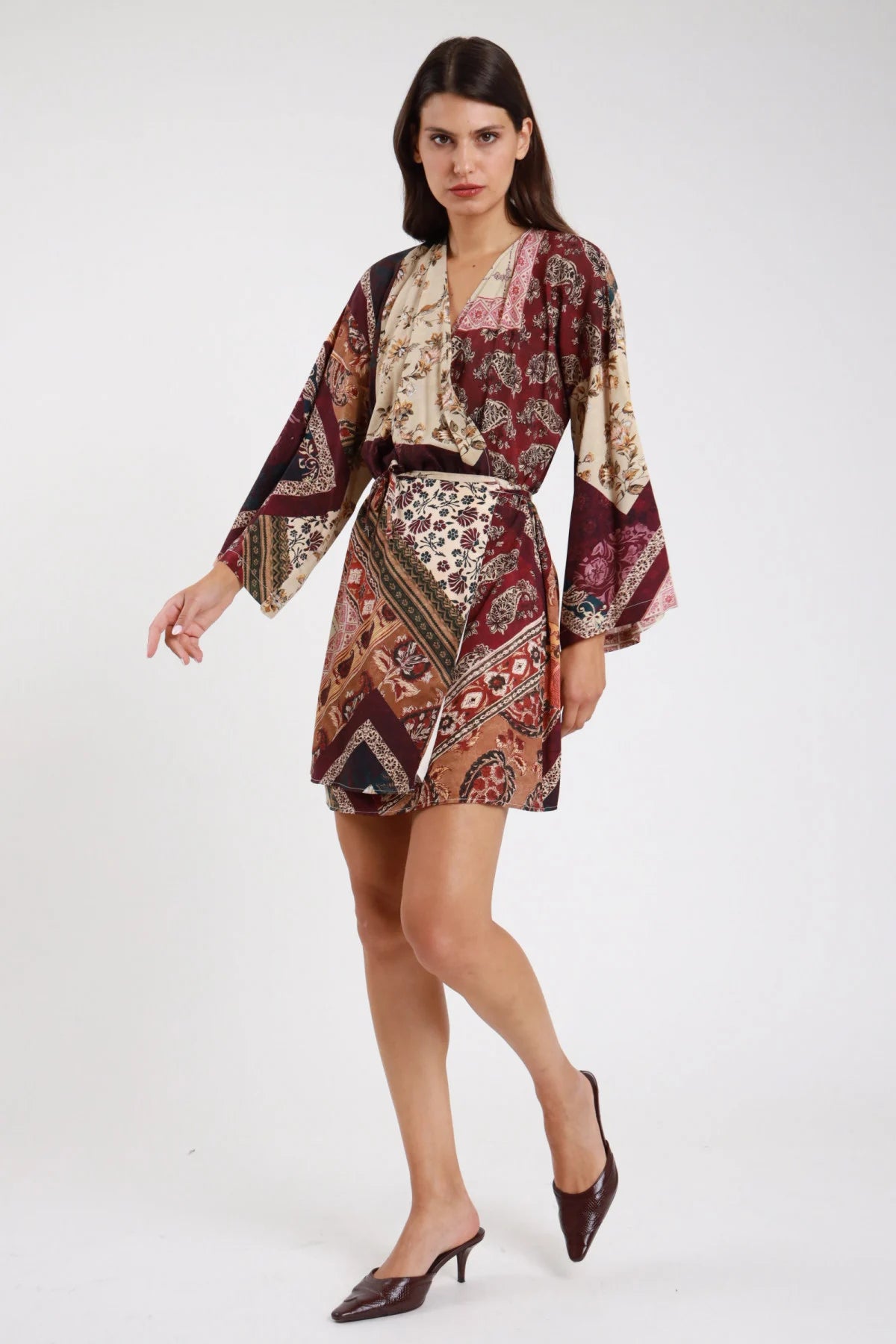 Abito kimono fantasia patchwork paisley baroque floral prugna bordeaux vinaccia panna ecru frontale intera tre quarti sguardo laterale scollo V laccetto fiocco drappeggiante manica kimono batwing ultralarga polsino campana flare svasato corpo oversize mini orlo asimmetrico cintura laccetto coordinato mules marroni tacco - modella frontale intera tre quarti sguardo laterale intenso mules marroni tacco capelli castani lisci sfondo bianco