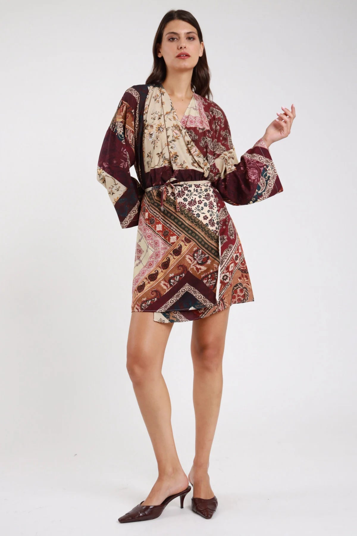 Abito kimono fantasia patchwork paisley baroque floral prugna bordeaux vinaccia panna ecru frontale intera mano laterale scollo V fiocco drappeggiante manica kimono batwing polsino campana flare corpo oversize mini orlo asimmetrico cintura laccetto nodo frontale mules marroni tacco - modella frontale intera mano laterale sguardo frontale intenso mules marroni tacco capelli castani lisci sfondo bianco