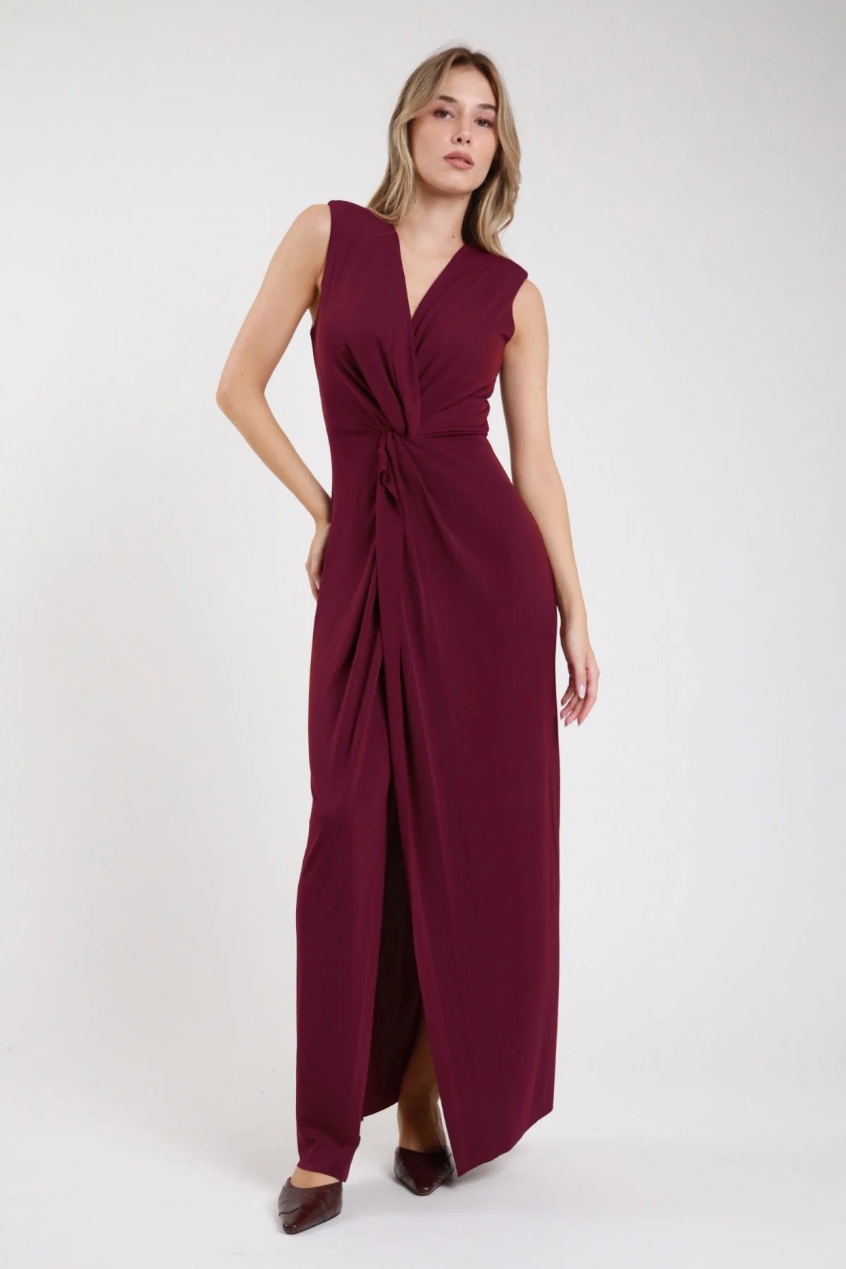 Abito maxi smanicato nodo bordeaux vino scollo V portafoglio drappeggio asimmetrico spacco - modella frontale intera braccio lungo fianco sguardo frontale tacchi marroni capelli biondi ondulati