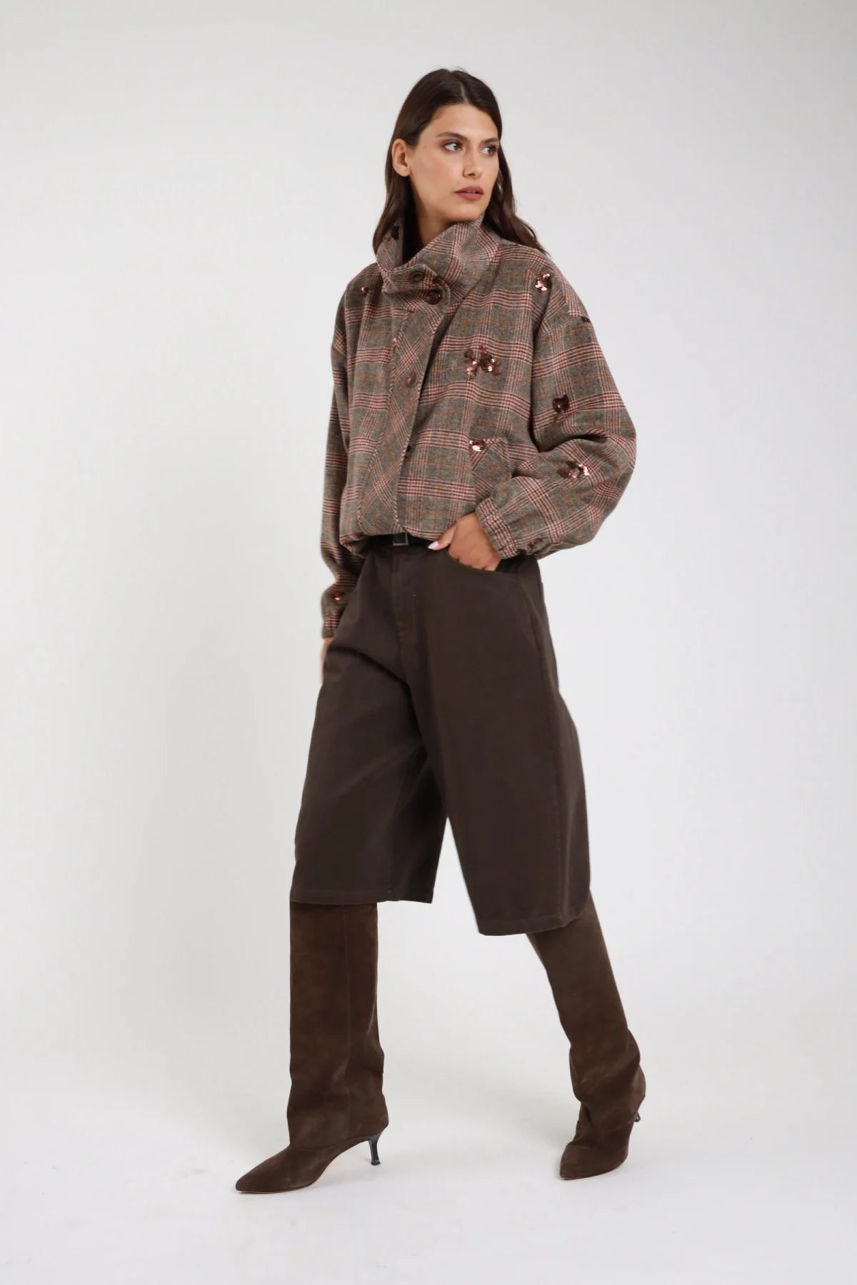 Bermuda culottes wide leg cioccolato moro scuro tre quarti laterale vita alta piega sartoriale cintura pelle nera fibbia argento tasche laterali gamba wide leg below-knee orlo dritto stivali marroni alti - modella tre quarti laterale mano tasca sguardo frontale con giacca bomber check tartan paillettes cipria stivali marroni alti capelli castani lisci sfondo bianco