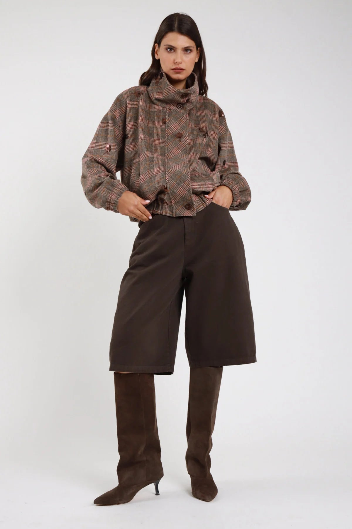 Bermuda culottes wide leg cioccolato moro scuro frontale intera vita alta piega sartoriale cintura pelle nera fibbia argento tasche laterali gamba wide leg below-knee orlo dritto stivali marroni alti - modella frontale intera mano tasca sguardo frontale con giacca bomber check tartan paillettes cipria stivali marroni alti capelli castani lisci sfondo bianco