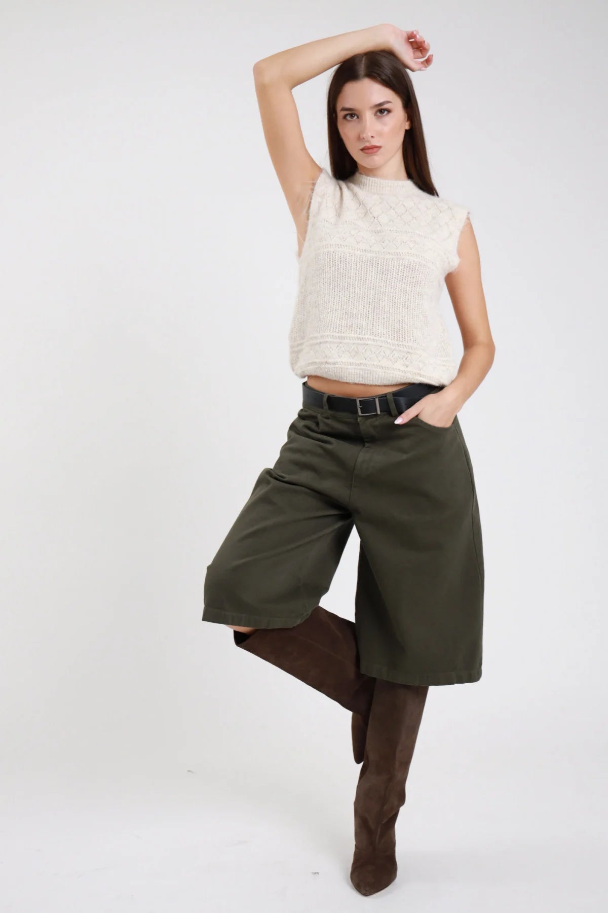 Bermuda culottes wide leg verde militare khaki frontale dinamica vita alta piega sartoriale cintura pelle nera fibbia argento tasche laterali gamba wide leg below-knee orlo dritto stivali marroni alti - modella frontale dinamica mano testa gamba alzata sguardo laterale alto con top knit panna mock neck stivali marroni alti capelli castani lisci sfondo bianco