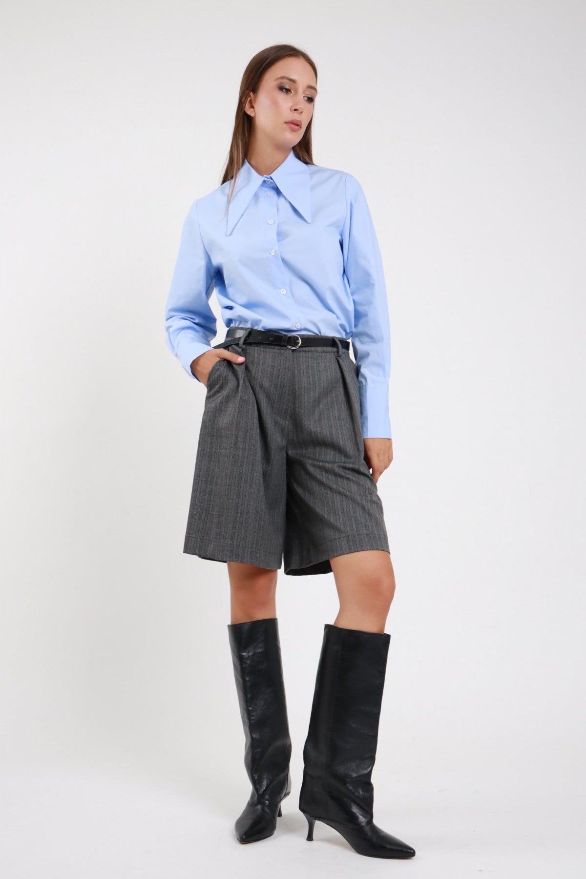 Bermuda Spinato herringbone grigio antracite lurex vita alta pince wide ginocchio frontale 3/4 abbinata camicia azzurra - modella frontale 3/4 mano tasca sguardo laterale capelli castani lisci sfondo bianco camicia azzurra collo classico cintura nera stivali neri tacco kitten