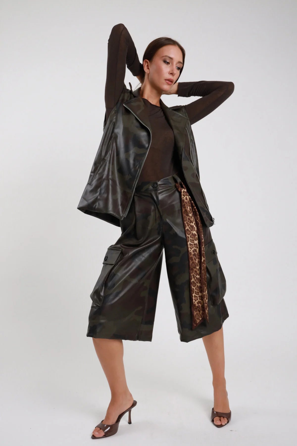 Bermuda Mimetico cargo culottes ecopelle PU camouflage verde bronzo tasconi laterali patta cintura leopardata vita alta frontale intera posa braccia alzate - modella frontale intera braccia alzate sguardo abbasso capelli castani lisci sfondo bianco giubbino mimetico aperto top marrone mules tacco marrone