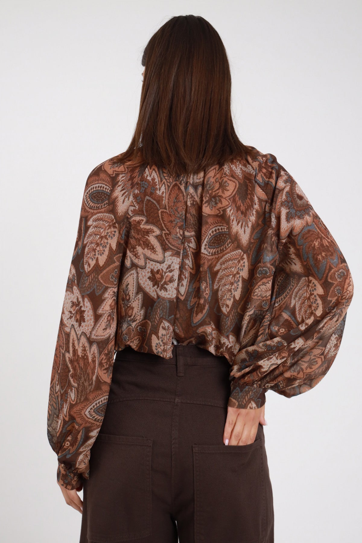 Blusa Lupetto Fantasia Paisley Cachemire chiffon marrone tabacco manica balloon voluminosa retro schiena silhouette oversize fluida - modella retro 3/4 sguardo abbasso capelli castani lisci sfondo bianco pantalone marrone scuro