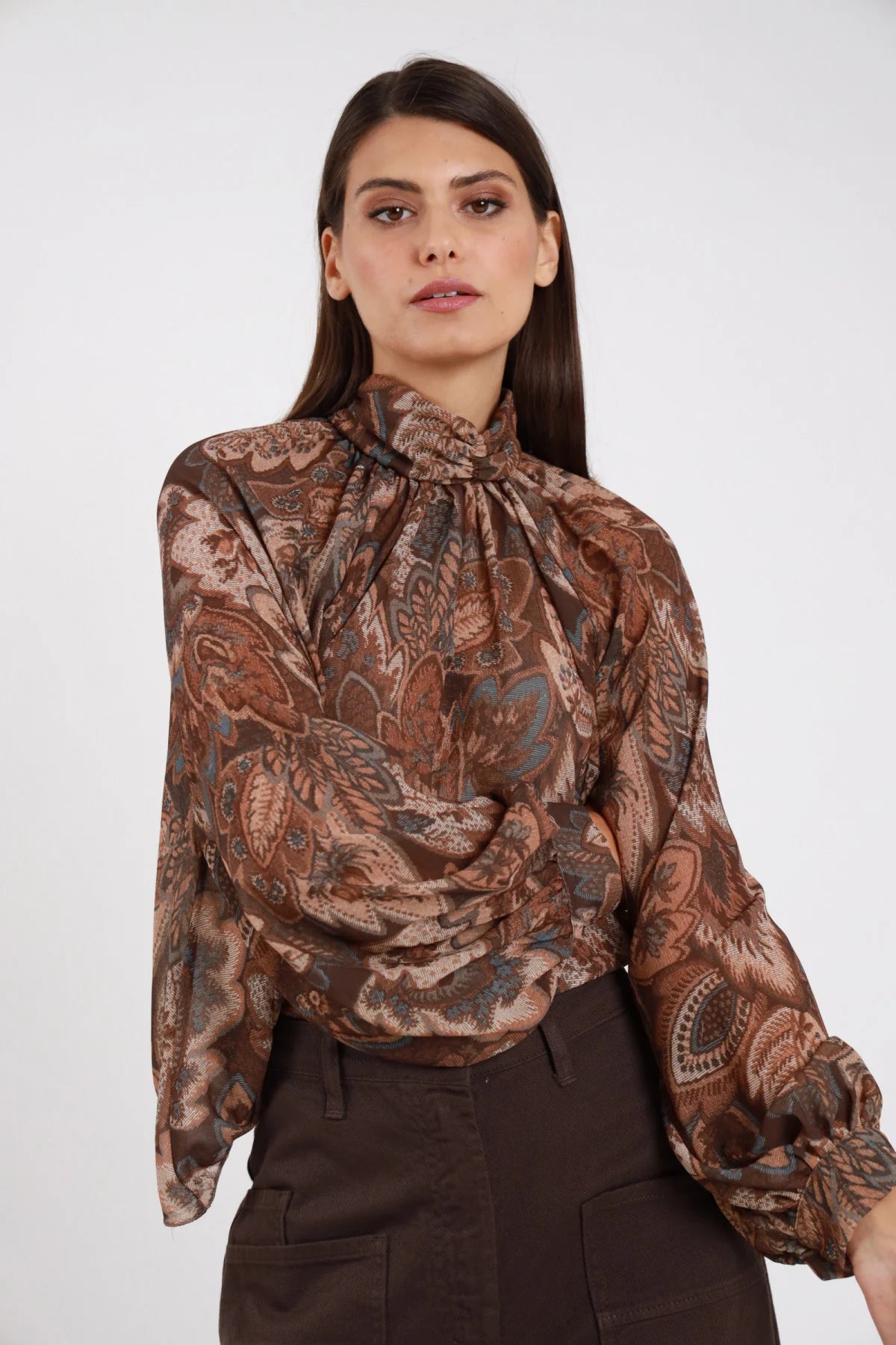 Blusa Lupetto Fantasia Paisley Cachemire chiffon marrone tabacco colletto lupetto arricciato coulisse manica balloon polsino elastico drappeggio frontale busto frontale 3/4 braccia incrociate - modella busto frontale 3/4 braccia incrociate sguardo intenso capelli castani lisci sfondo bianco pantalone marrone scuro