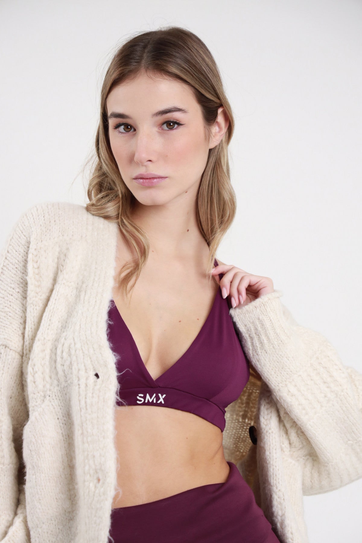 Bralette SMX Limited Edition bordeaux prugna 95% tactel triangolo scollo V fascia elastica logo SMX bianco crop busto frontale cardigan panna chunky aperto mano colletto - modella busto frontale cardigan panna aperto mano colletto sguardo frontale capelli biondi mossi sfondo bianco gonna bordeaux abbinata