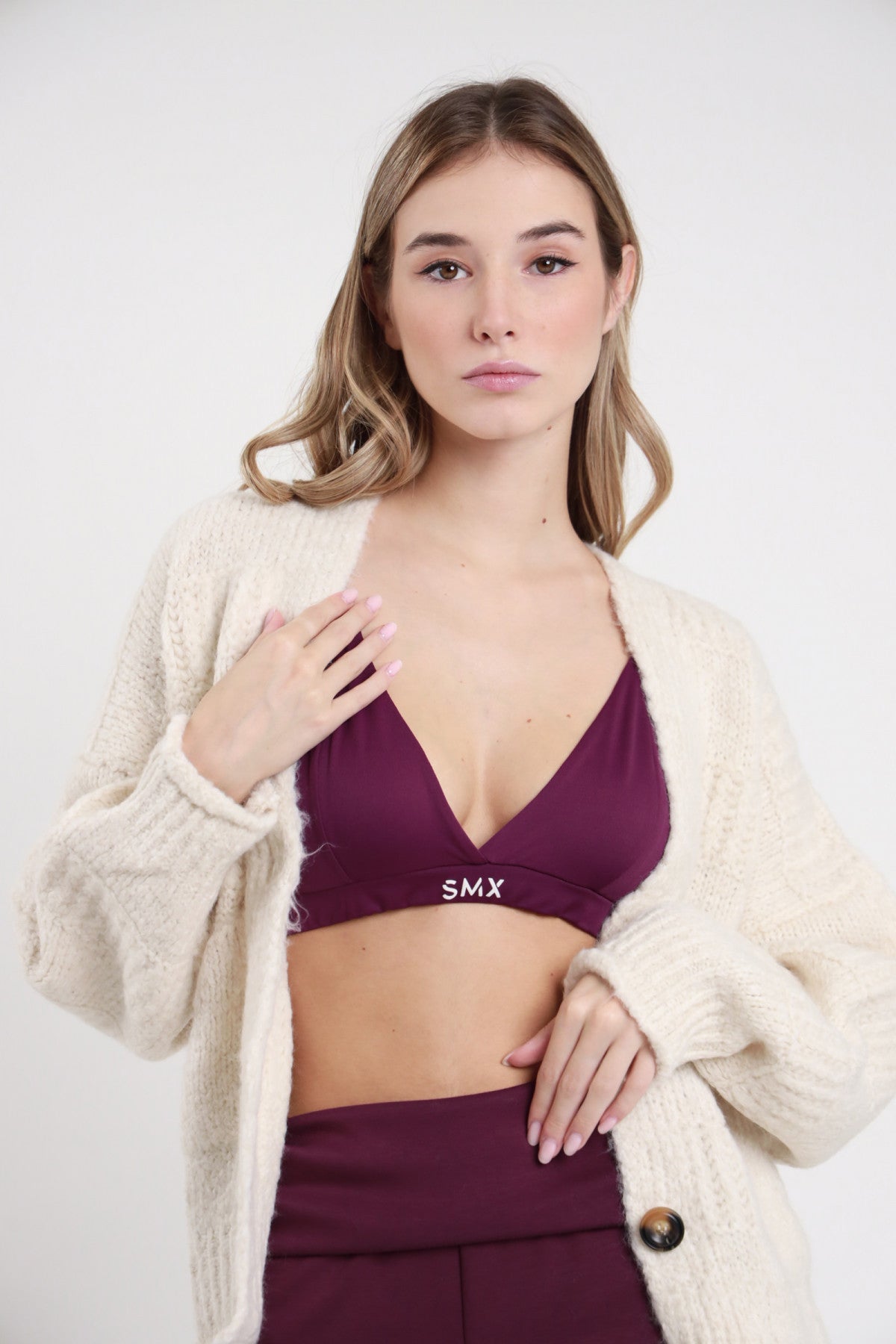 Bralette SMX Limited Edition bordeaux 95% tactel triangolo scollo V fascia elastica logo SMX bianco crop busto 3/4 cardigan panna aperto mano petto - modella busto 3/4 cardigan panna aperto mano petto sguardo frontale capelli biondi mossi sfondo bianco gonna bordeaux abbinata