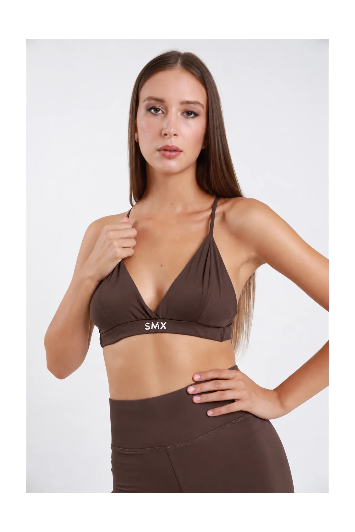 Bralette Triangolo DAYTHRU marrone cioccolato scuro scollo V profondo incrociato spalline sottili fascia elastica logo SMX bianco a contrasto second-skin busto frontale 3/4 posa mano spallina sguardo frontale - modella busto frontale 3/4 posa mano spallina sguardo frontale capelli castani lisci sfondo bianco leggings marrone coordinati