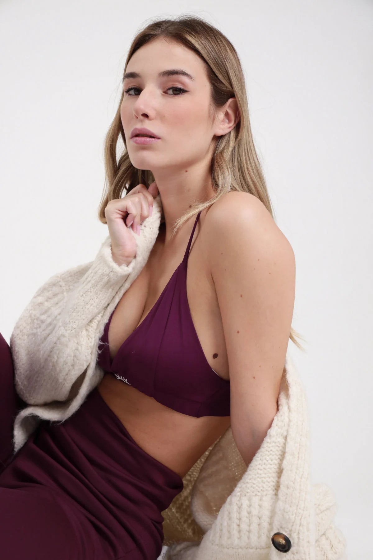 Bralette SMX Limited Edition bordeaux prugna 95% tactel triangolo spalline incrociate cross-back fascia logo SMX crop seduta 3/4 laterale cardigan panna spalla sguardo alto - modella seduta 3/4 laterale cardigan panna sulla spalla sguardo alto capelli biondi sciolti sfondo bianco gonna bordeaux abbinata