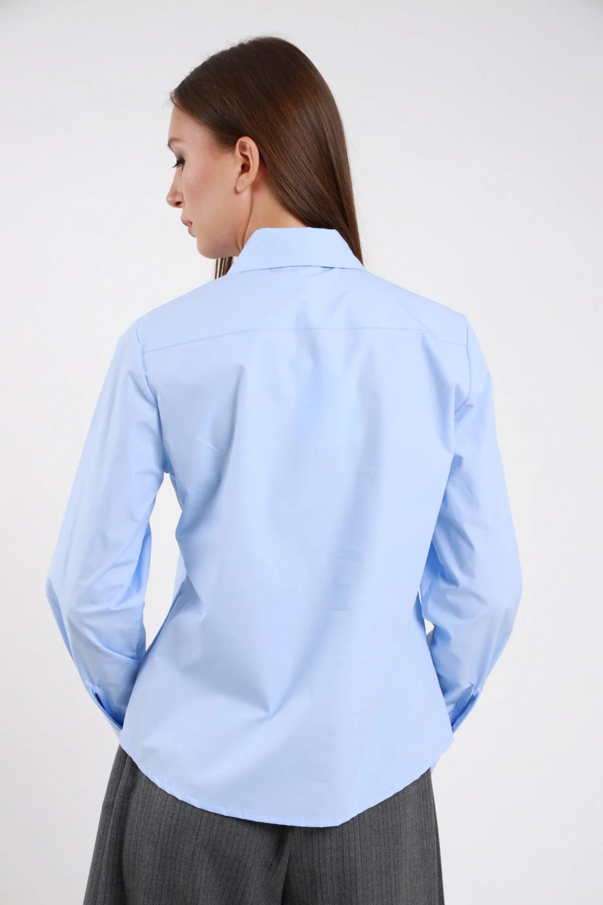 Camicia Oversize Maxi Colletto Appuntito azzurro celeste 100% cotone popeline posteriore silhouette oversize fondo curvo arrotondato colletto posteriore visibile - modella intera posteriore posa statica mani fianchi sguardo laterale capelli castani lisci sfondo bianco pantalone wide leg grigio