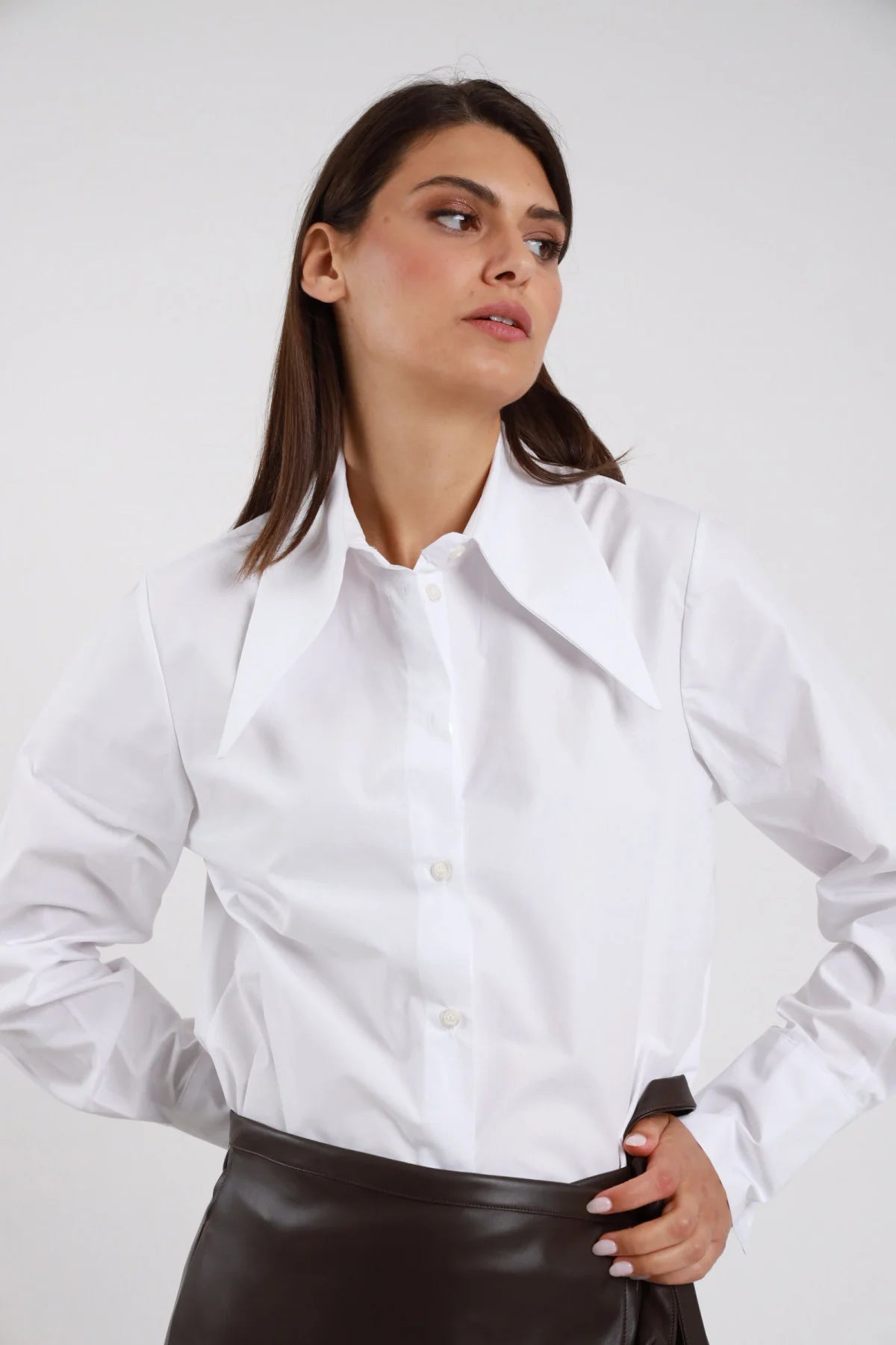 Camicia Oversize Maxi Colletto Appuntito bianco 100% cotone popeline maxi colletto punta lunga strutturato bottoni bianchi ton-sur-ton polsino bottone silhouette oversize morbida - modella busto frontale 3/4 posa mani fianchi sguardo laterale capelli castani lisci sfondo bianco gonna minigonna ecopelle marrone scuro