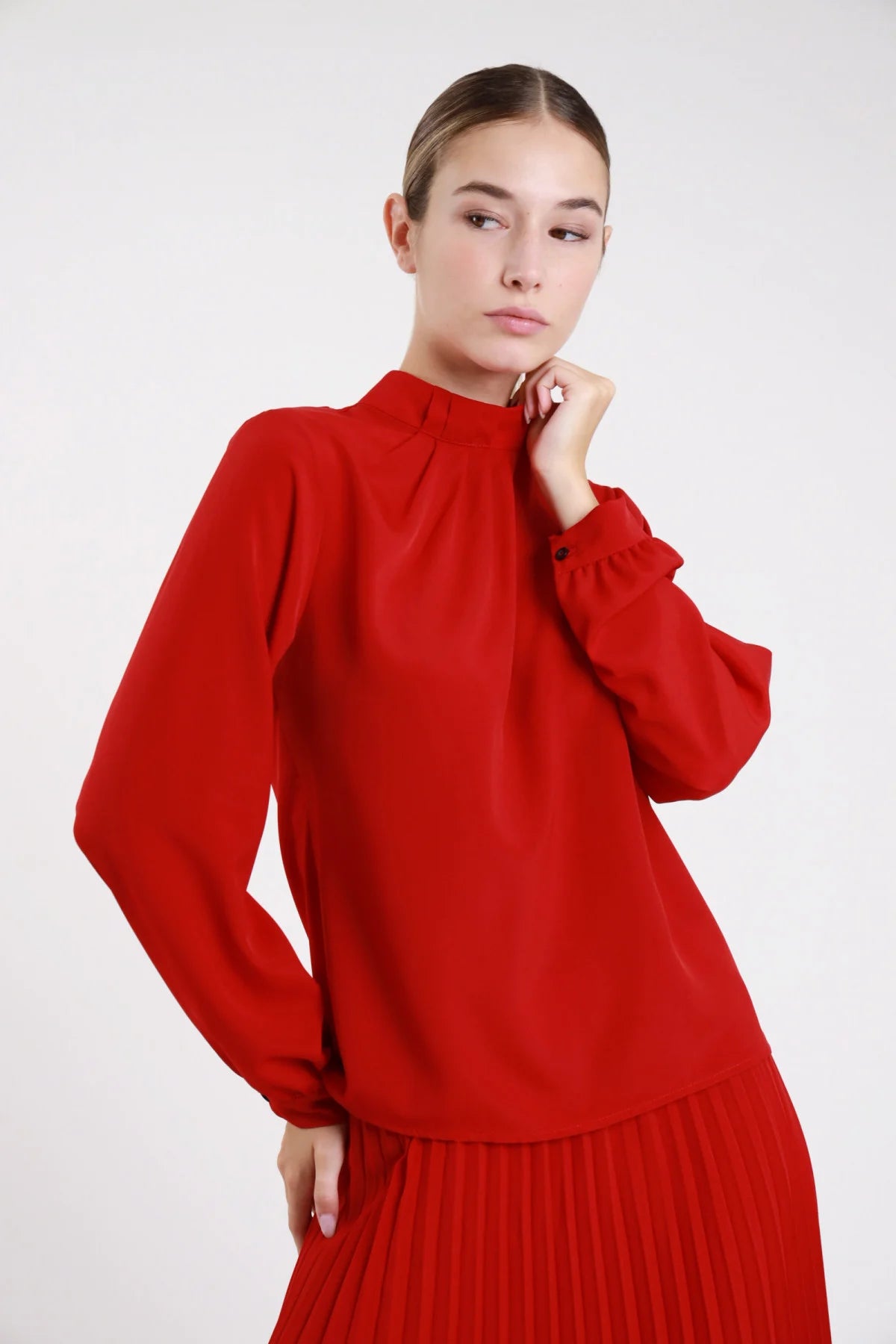 Camicia pieghe collo alto fluida rossa maniche palloncino - modella busto tre quarti mano al mento sguardo laterale con gonna plissettata rossa abbinata capelli castani lisci