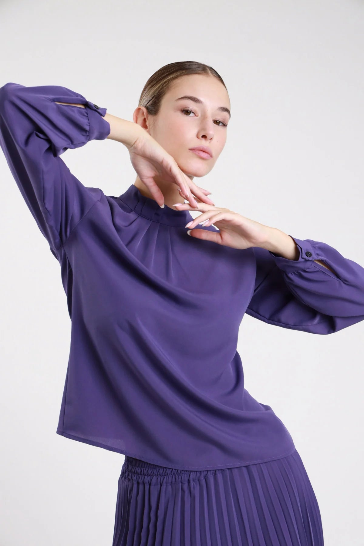 Camicia pieghe collo alto fluida viola indaco arricciatura palloncino - modella busto tre quarti braccia alzate mani al collo gonna plissettata viola capelli castani raccolti