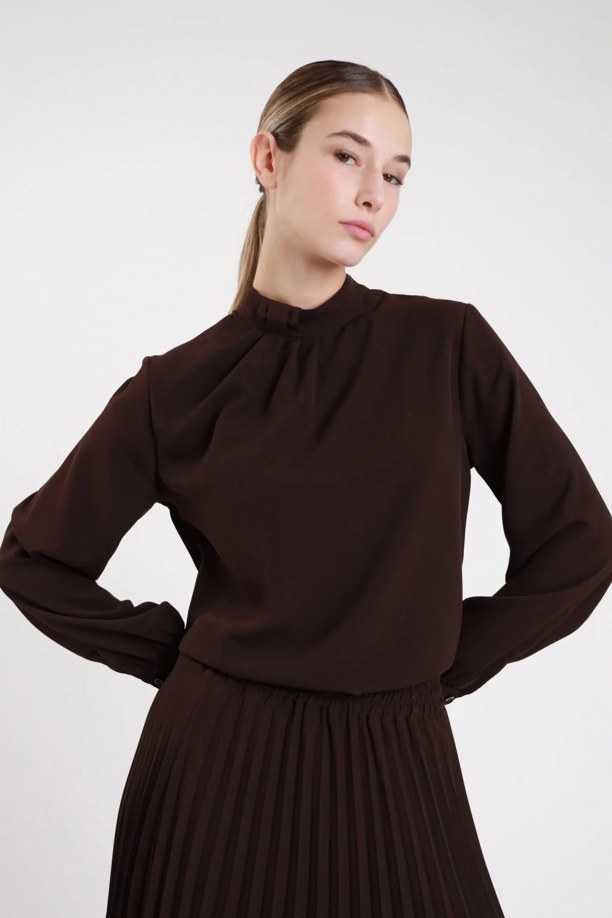 Camicia pieghe collo alto fluida cioccolato scuro taglio ampio - modella busto frontale mani sui fianchi sguardo frontale gonna plissettata cioccolato capelli castani coda