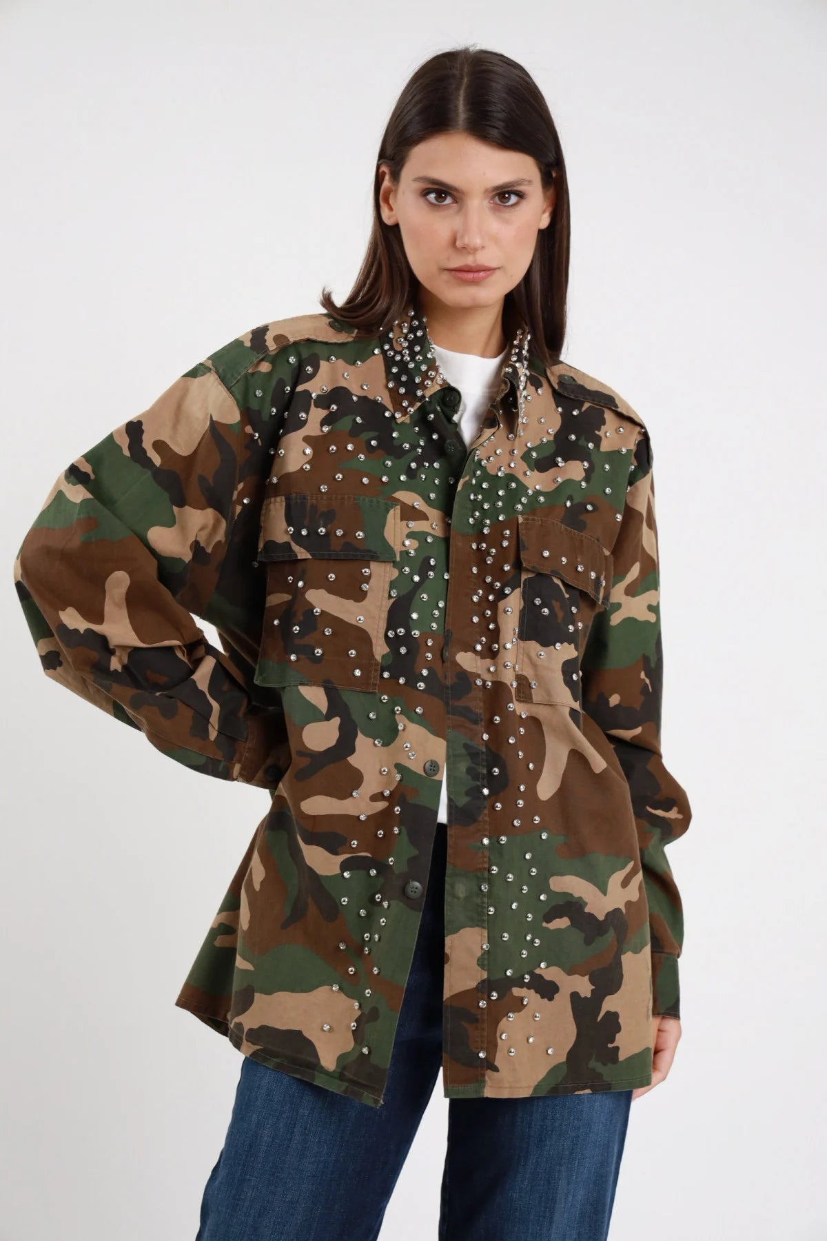Camicia Militare Mimetica Woodland Borchie Strass 95% cotone aperta t-shirt bianca sotto colletto borchiato aperto strass argentati pioggia davanti tasche silhouette oversize lunga maxi frontale - modella busto frontale mano fianco sguardo frontale intenso capelli castani lisci sfondo bianco jeans denim scuro