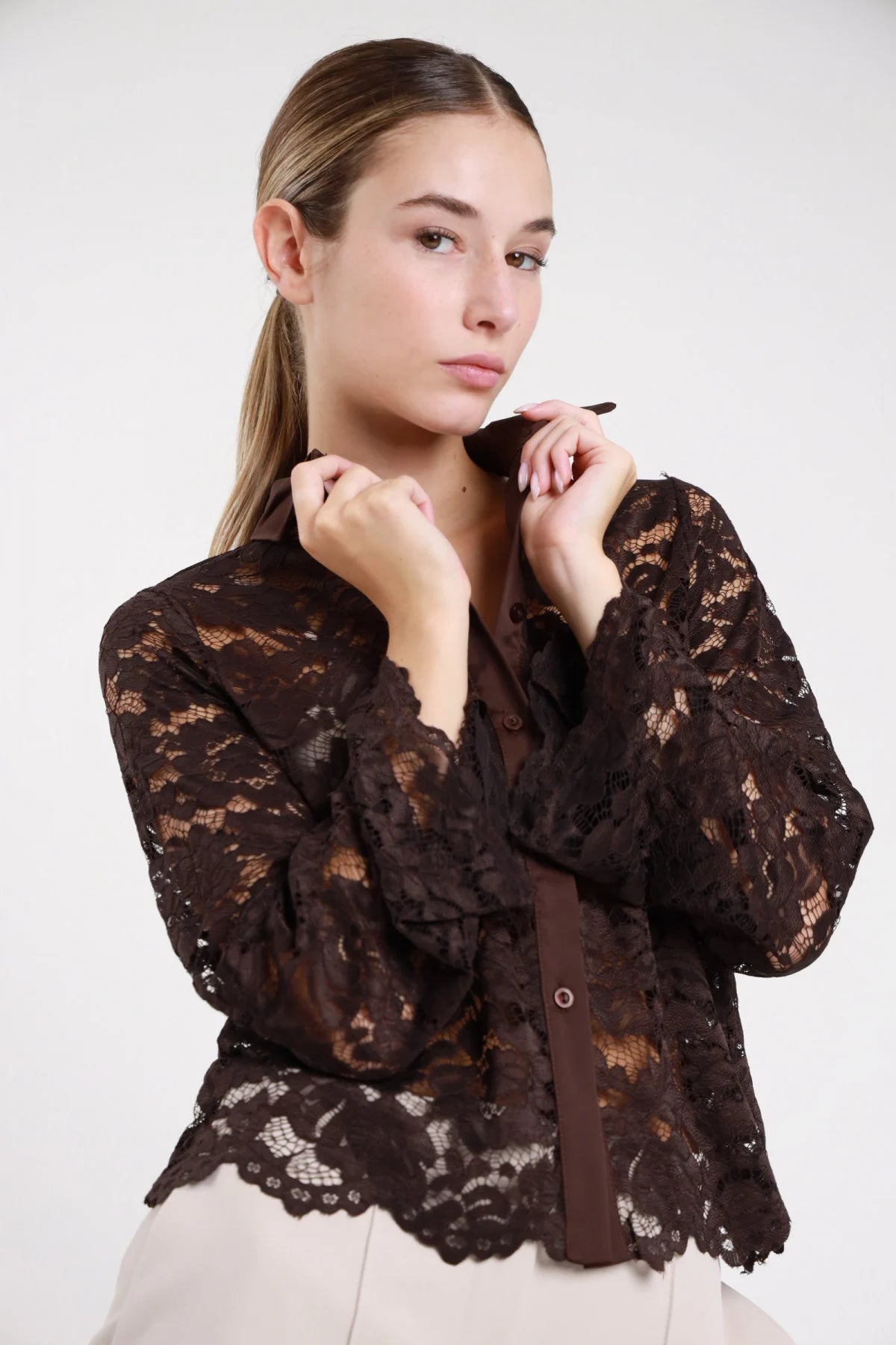 Camicia pizzo cioccolato moro scuro semitrasparente frontale tre quarti mani colletto sguardo basso manica campana flare orlo scallop smerlato floreale bottoni cioccolato - modella frontale tre quarti mani colletto sguardo basso pantaloni beige capelli biondi lisci raccolti