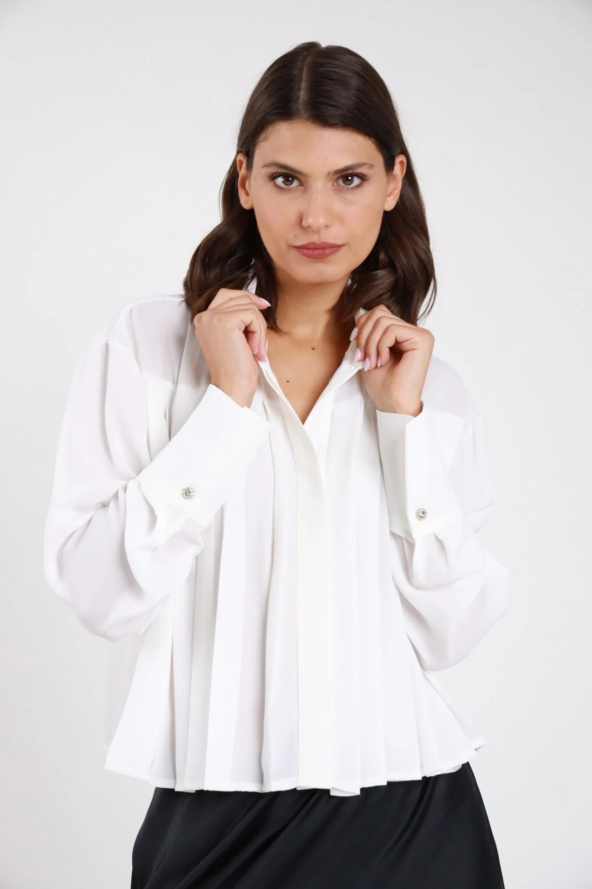 Camicia plissé bianca panna voile crepe fluido pince verticali frontali colletto classico scollo V manica lunga polsino doppio bottone gioiello strass oversize crop frontale busto mani colletto - modella frontale busto mani colletto sguardo frontale capelli castani mossi medi sfondo bianco gonna satin nera