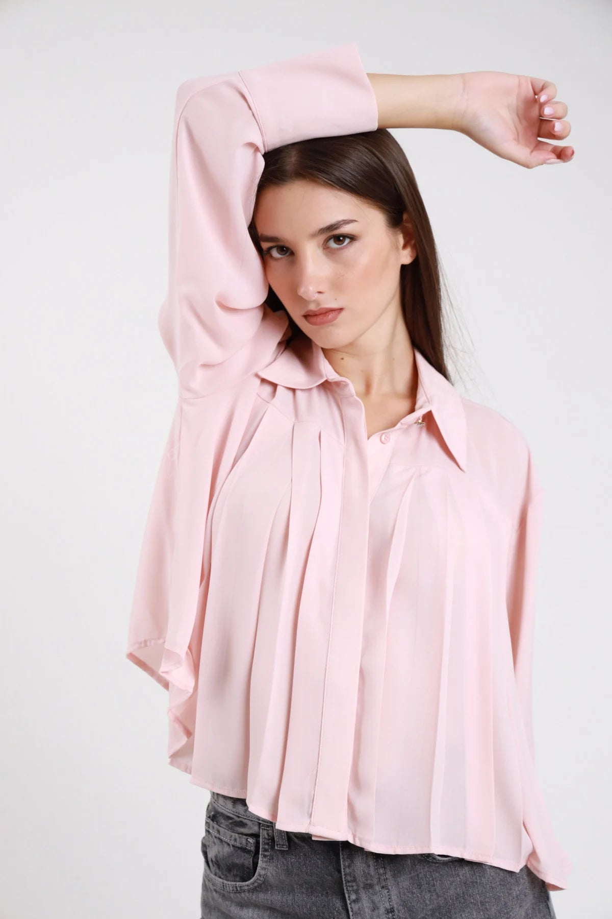 Camicia plissé rosa cipria chiaro voile crepe fluido pince verticali frontali colletto classico scollo V manica lunga polsino doppio bottone gioiello oversize crop frontale busto braccio alzato - modella frontale busto braccio alzato sguardo laterale capelli castani lisci sfondo bianco jeans grigio scuro