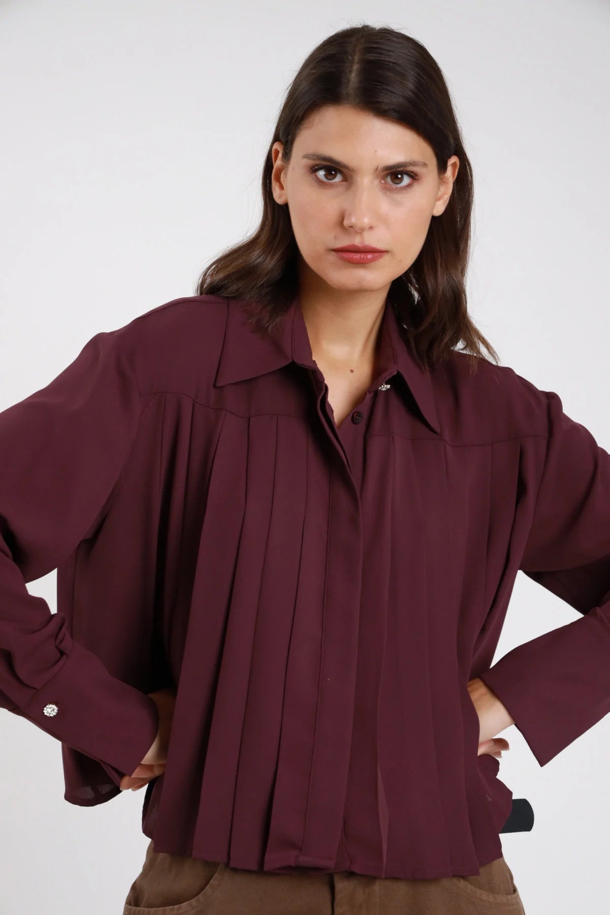 Camicia plissé bordeaux vinaccia scuro voile crepe fluido pince verticali frontali colletto classico scollo V manica lunga polsino doppio bottone gioiello strass oversize crop frontale busto mani fianchi - modella frontale busto mani fianchi sguardo frontale intenso capelli castani scuri lisci sfondo bianco pantaloni cammello