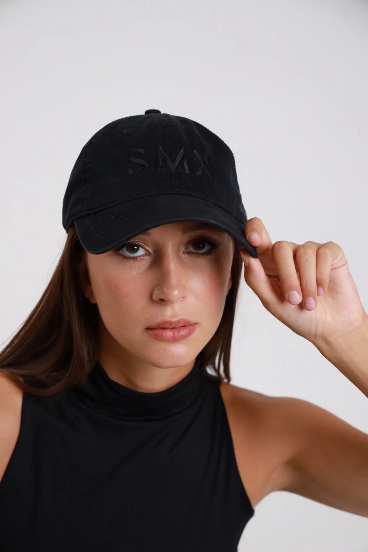 Cappellino Baseball DAYTHRU nero logo SMX ricamato tono su tono nero calotta frontale visiera curva strutturata dad cap - modella primo piano frontale mano che regge visiera sguardo intenso frontale capelli castani lisci sfondo bianco maglia collo alto nera senza maniche