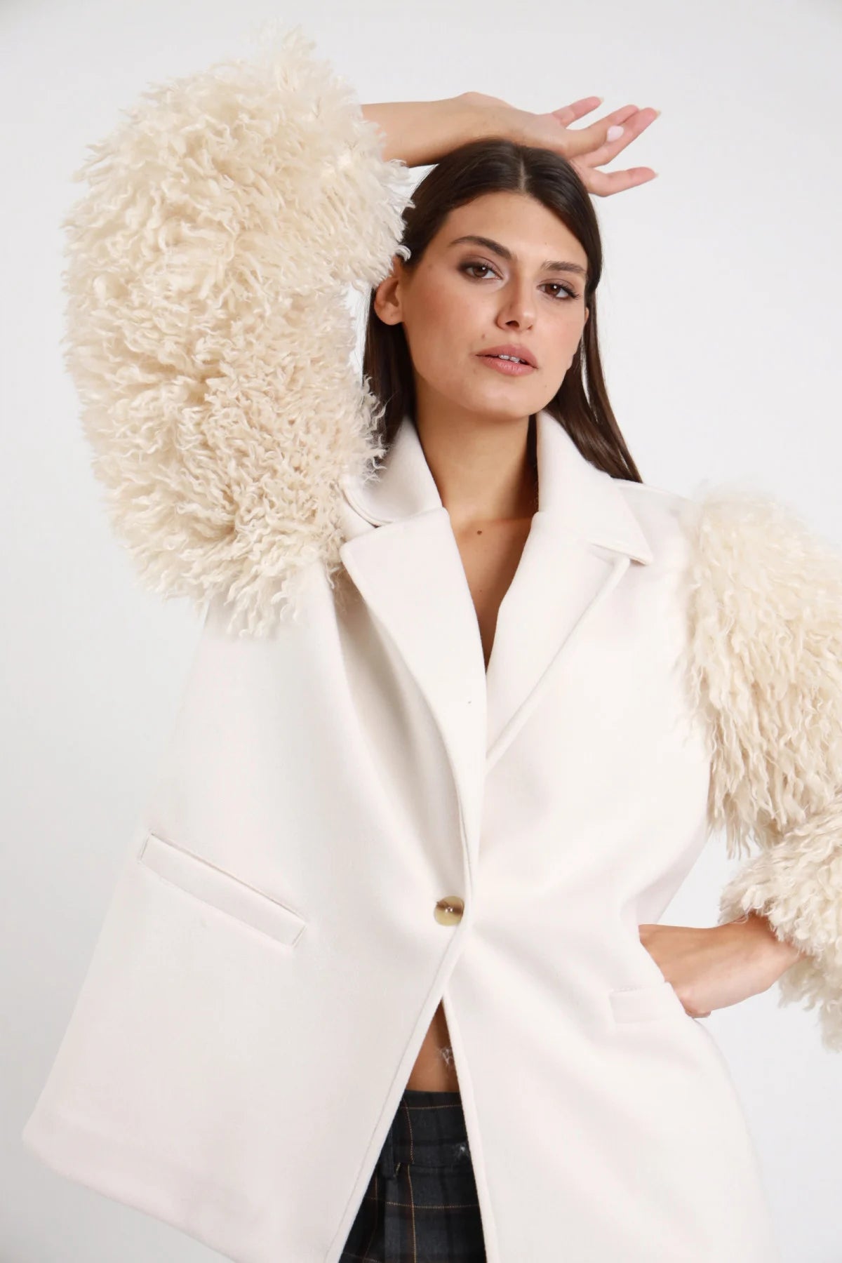 Blazer Maniche Ecopelliccia panna avorio 100% poliestere revers V bottone dorato tascone pattina manica ecopelliccia panna alzata volume riccioluto enorme vaporoso dettaglio busto braccio alzato posa dinamica - modella busto 3/4 braccio alzato posa dinamica sguardo frontale capelli castani lisci sfondo bianco pantalone tartan grigio abbinato