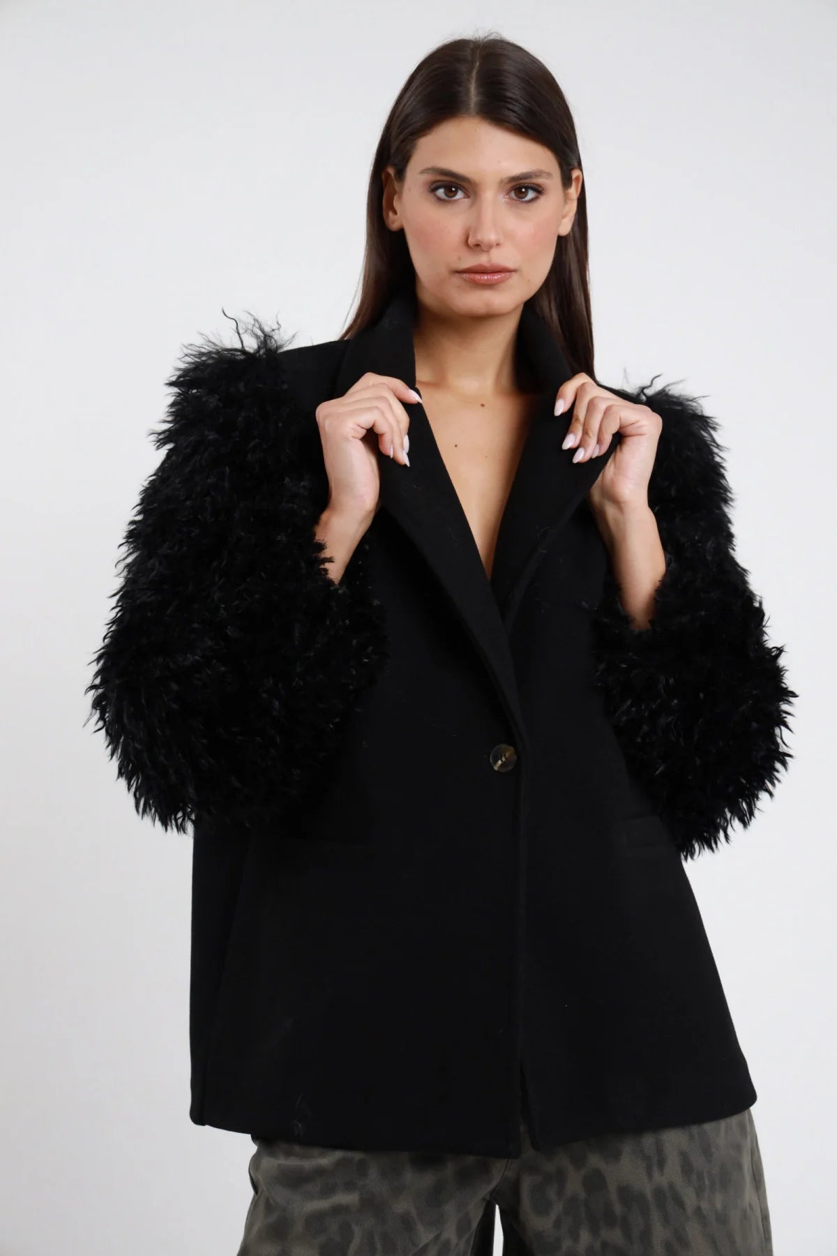 Blazer Maniche Ecopelliccia nero 100% poliestere doubleface revers V sartoriale 1 bottone nero tasche pattina maniche ecopelliccia nera riccioluta sintetica voluminose drammatiche silhouette oversize frontale busto mani revers - modella busto frontale mani revers sguardo frontale capelli castani lisci sfondo bianco pantaloni animalier leopardati abbinati