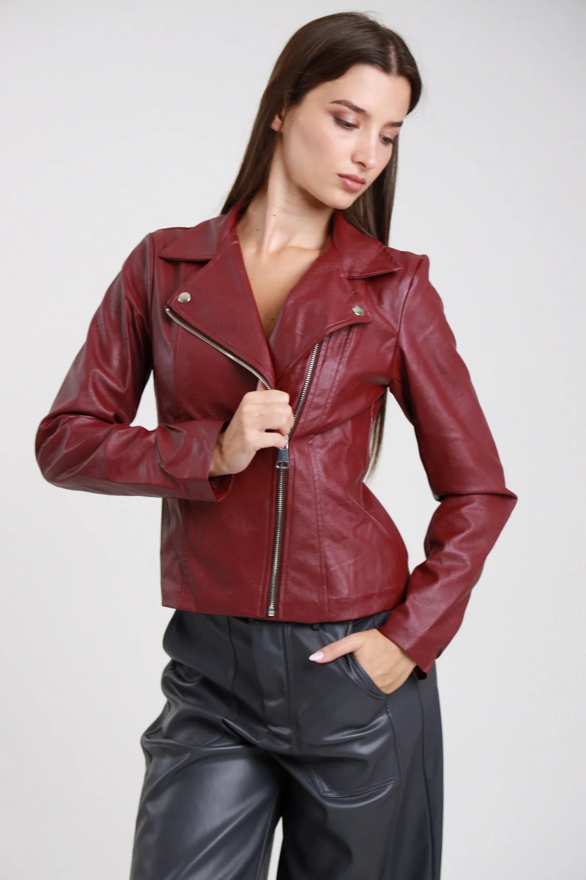 Chiodo ecopelle bordeaux vinaccia donna slim cropped colletto revers zip frontale asimmetrica argento tasche laterali zip - vista frontale tre quarti mano alla zip con pantaloni ecopelle grigi